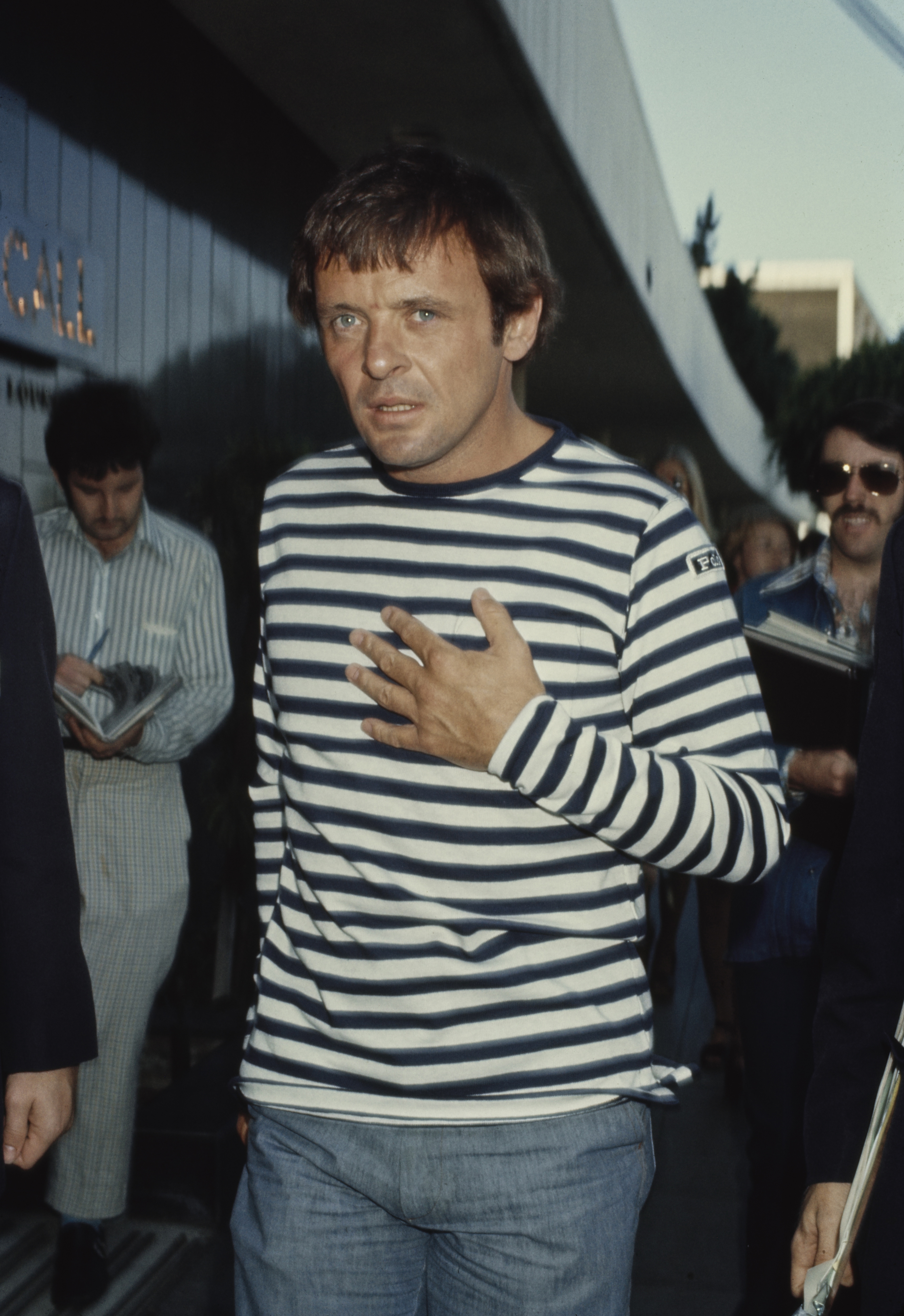 Anthony Hopkins strides purposefully as his expression is caught between thought and intensity. Dressed in a casual navy-and-white striped shirt, he exudes effortless charm with a touch of mystery. The fading daylight and onlookers around him lend the scene a cinematic sense of quiet anticipation.
