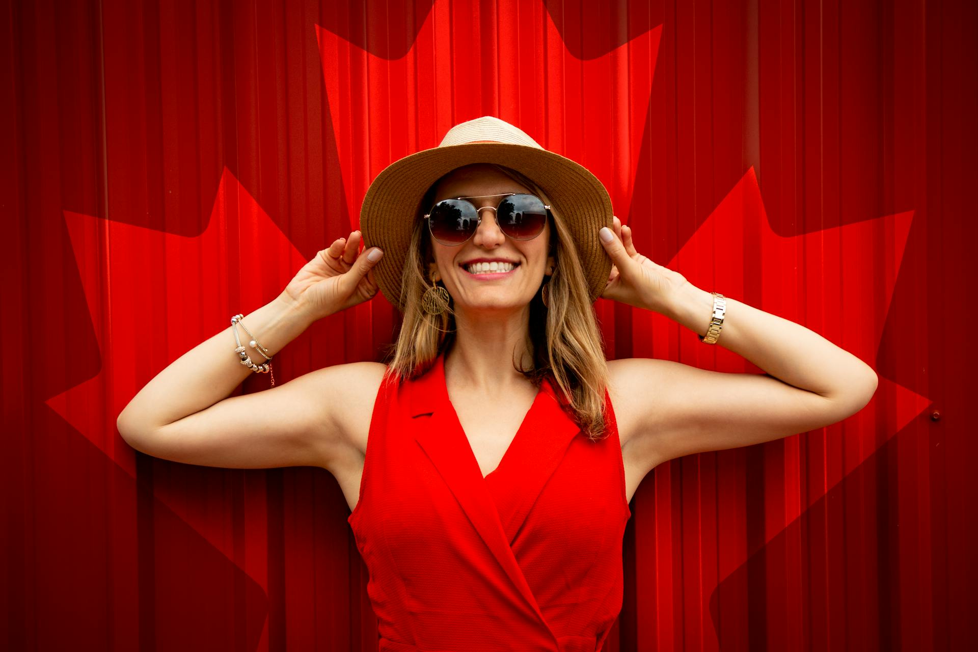 A smiling woman in a red dress, sunglasses, and a hat standing against a red backdrop | Source: Pexels