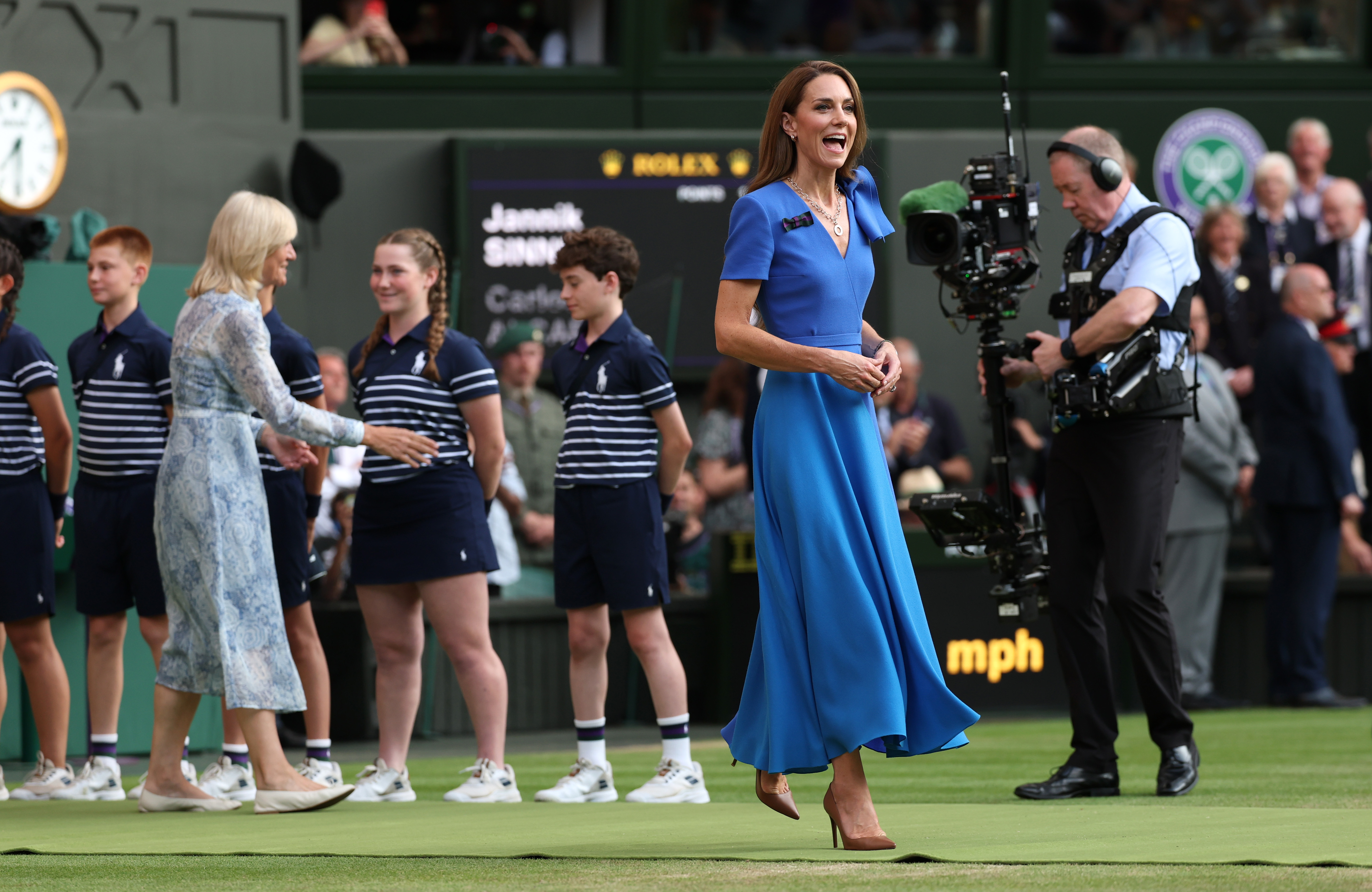Princess Catherine makes her way toward the trophy table, walking past the lined-up ball boys and girls. She wears a calm expression as cameras follow her movement on the grass court.