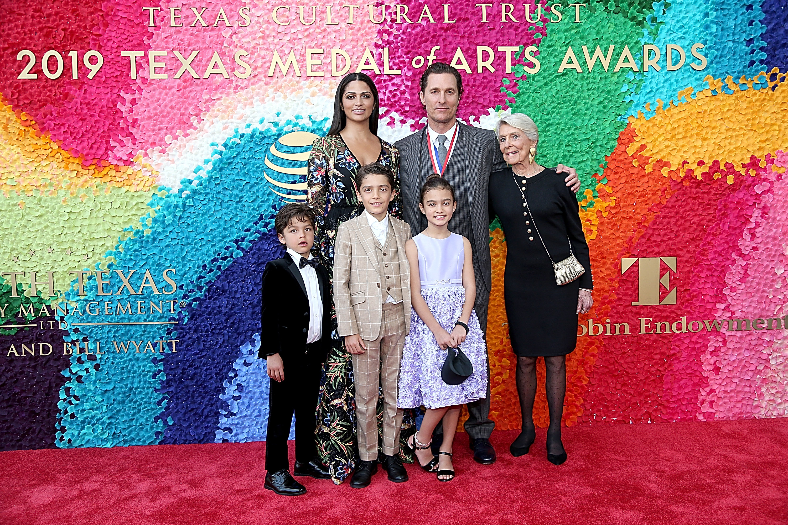 Levi, Vida, and Livingston Alves McConaughey stand in front in formal outfits. Levi wears a checked suit, Vida wears a light dress with a small bag, and Livingston wears a dark suit. Matthew McConaughey, Camila Alves, and Kay McConaughey stand behind them on the red carpet.
