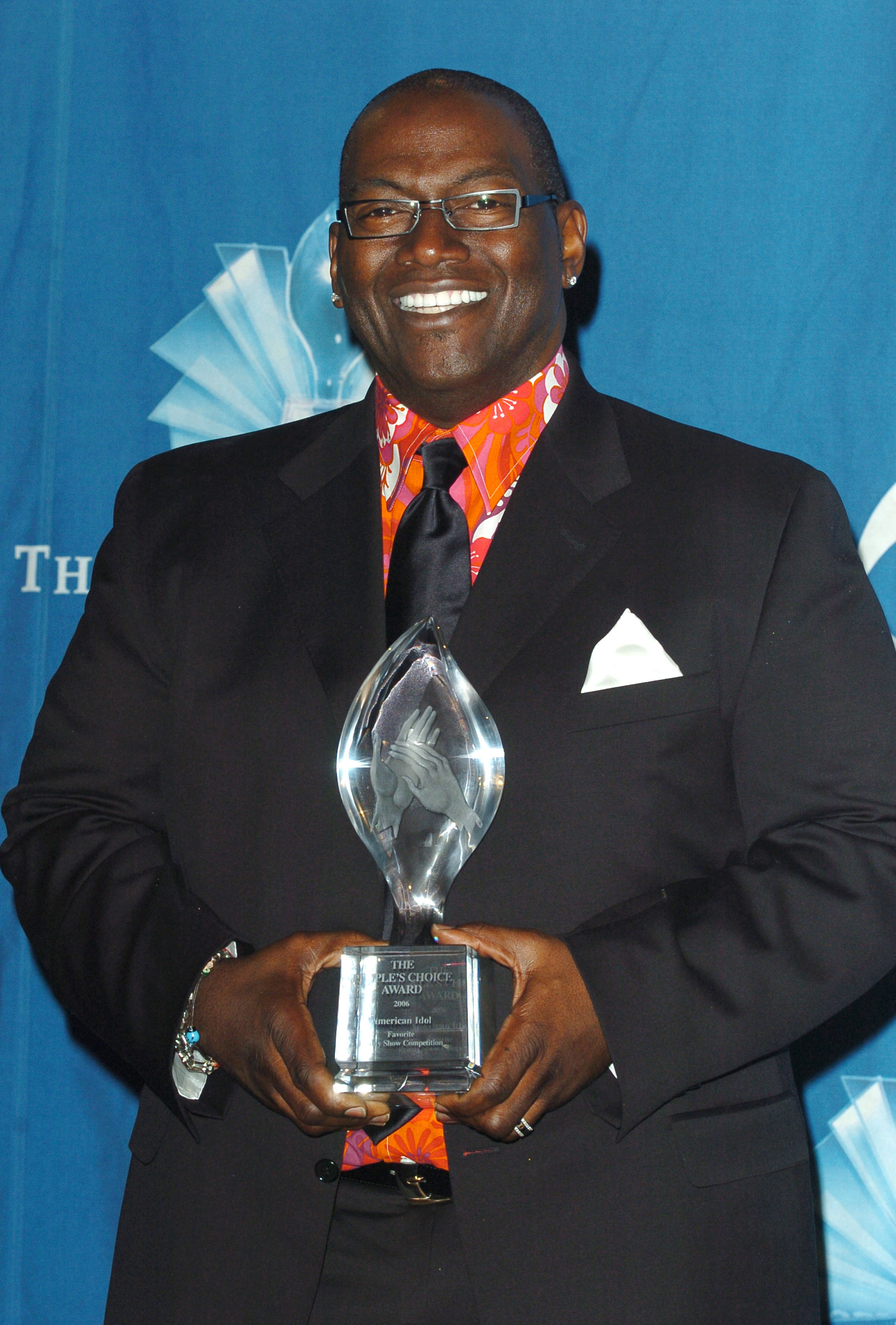 Holding a crystal People’s Choice Award, Randy Jackson beams in a sharp black suit layered over a vibrant patterned shirt and satin tie.