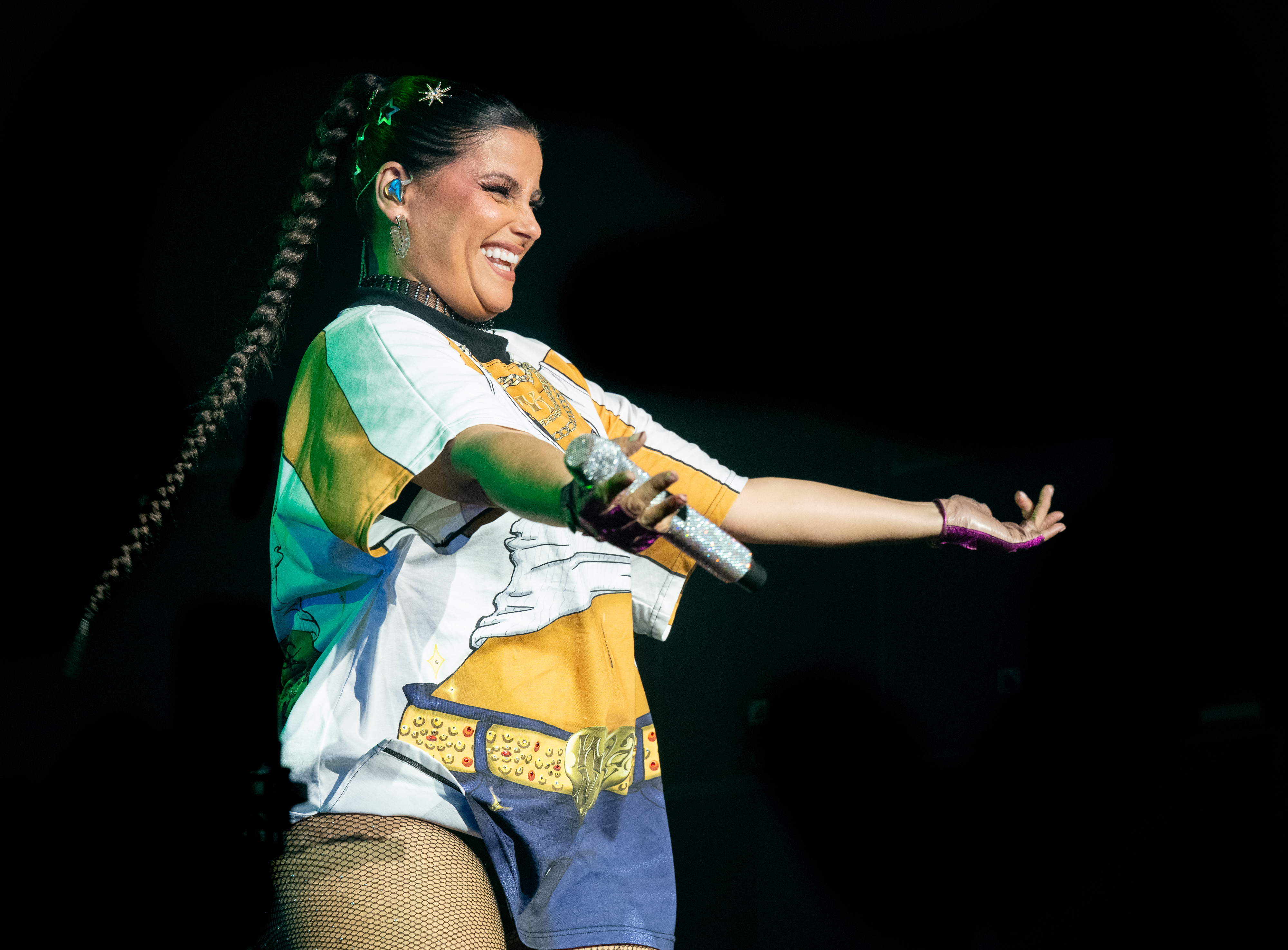 Furtado performs on stage in a graphic t-shirt with a belt-inspired design, paired with shorts and fishnet stockings. Her hair is styled in a long braid, and she accessorizes with earrings, a choker necklace, and bold makeup. She holds a microphone in one hand and gestures to the crowd with the other.