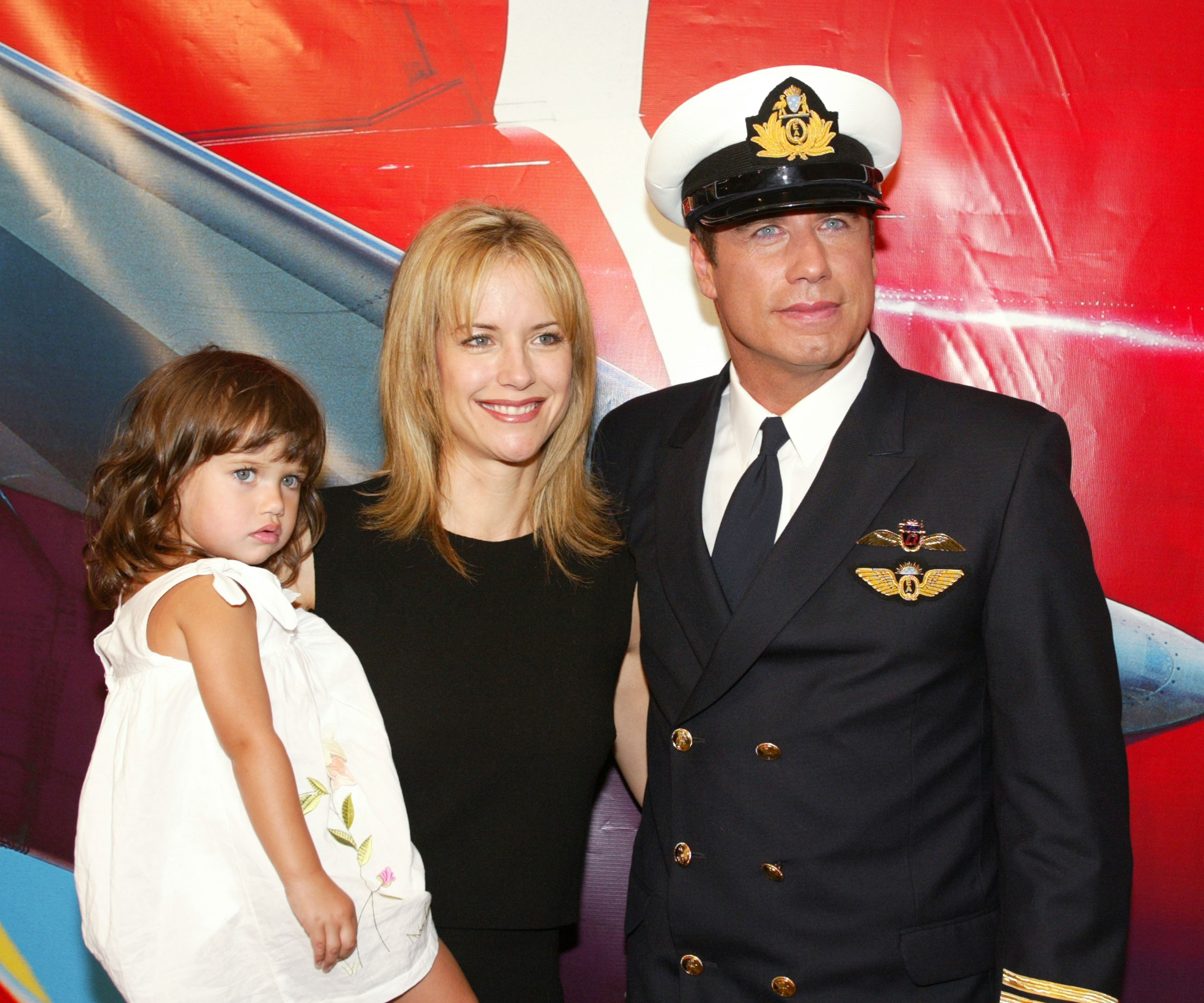A young Ella Bleu Travolta is held by her mom, Kelly Preston, as John Travolta stands beside them in a pilot uniform at LAX. The family appeared at a 2002 press event announcing John's role as a Qantas ambassador.