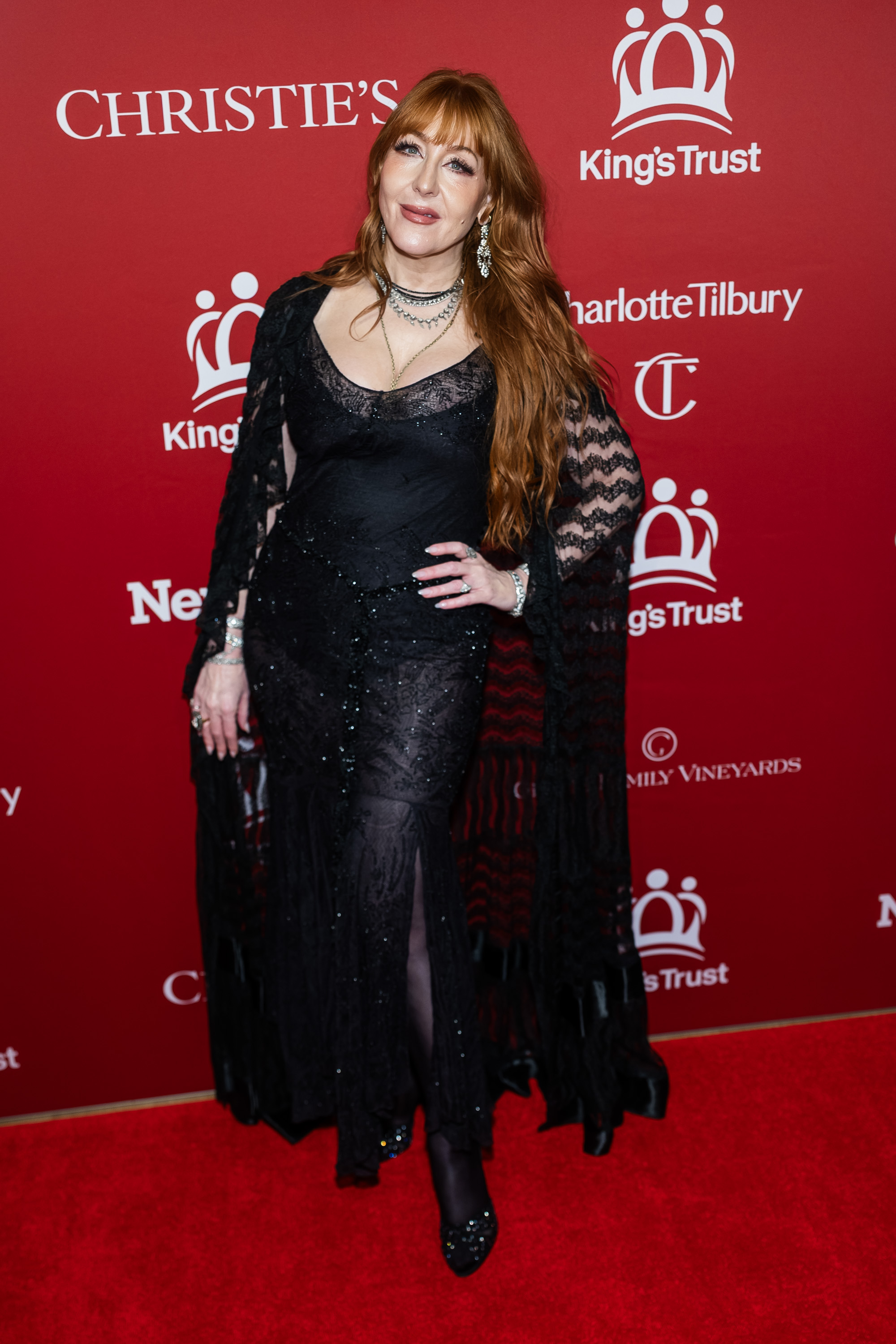 Charlotte Tilbury works the indoor step-and-repeat in a black lace semi-sheer gown scattered with tiny crystals, paired with a dramatic floor-length lace cape, layered silver chain necklaces, chandelier earrings, and sparkling embellished heels, her auburn waves cascading freely.