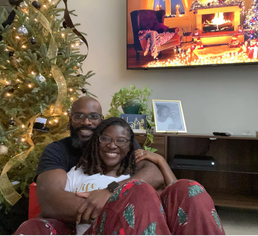 Nancy Metayer Bowen sits on the floor in cozy holiday pajamas, smiling as her husband, Stephen Bowen, wraps his arms around her from behind, with a decorated Christmas tree and a warmly lit living room creating an intimate festive atmosphere. | Source: Instagram/nancymetayerbowen