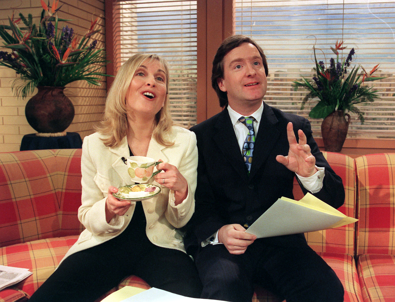 In a second shot from the same 1998 broadcast, Fiona Phillips was photographed once more on the GMTV sofa with her husband and reporter Martin Frizell, continuing the light-hearted exchange seen moments earlier, with the image capturing a more animated moment that reinforced their ease together on live morning television.