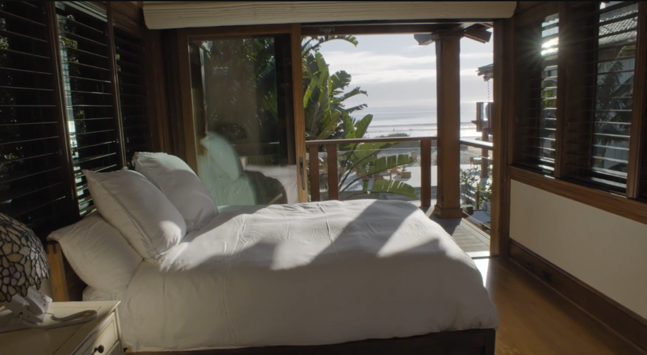 Sliding glass doors in a tranquil guest bedroom pull back to reveal a private wooden balcony with sweeping views of the Pacific Ocean. Crisp white linens cover the bed, while dark wood window shutters and warm flooring enhance the tropical, serene ambiance. | Source: YouTube/Architectural Digest