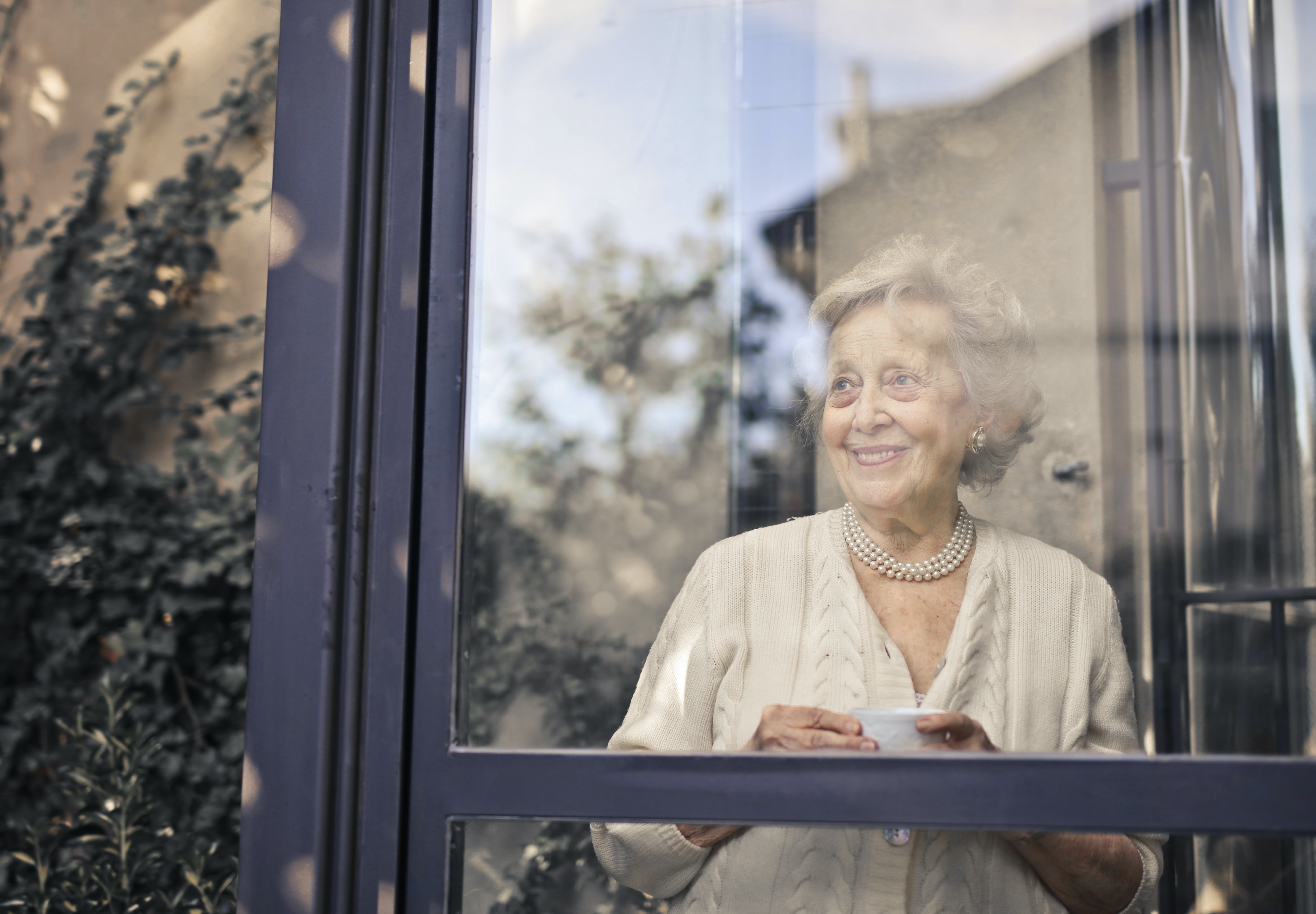 An older woman smiling while holding a beverage and looking out of a window | Source: Pexels