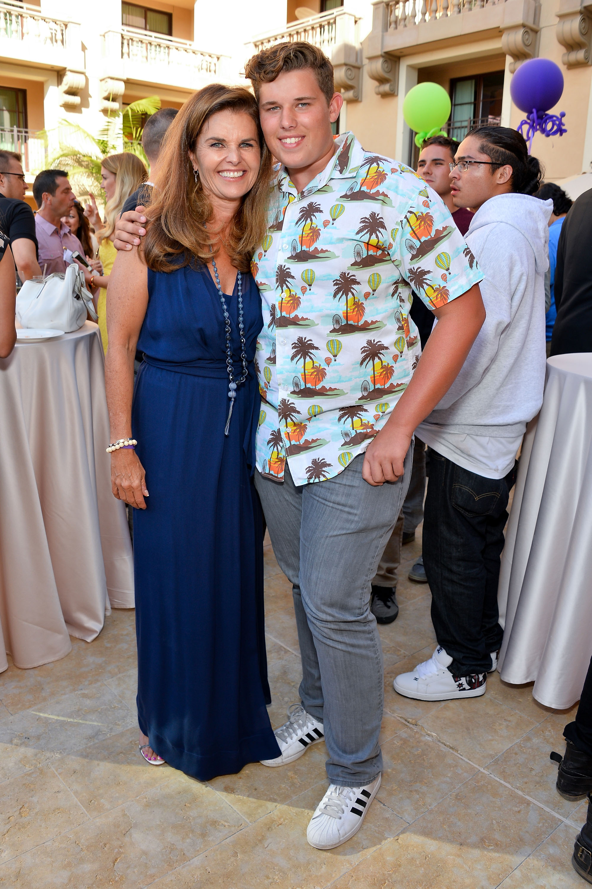 Maria Shriver and her son Christopher share a warm moment at the same event. Shriver wears a deep blue dress with layered necklaces, while Christopher sports his signature printed shirt and grey denim. They stand close, surrounded by guests in a festive atmosphere.