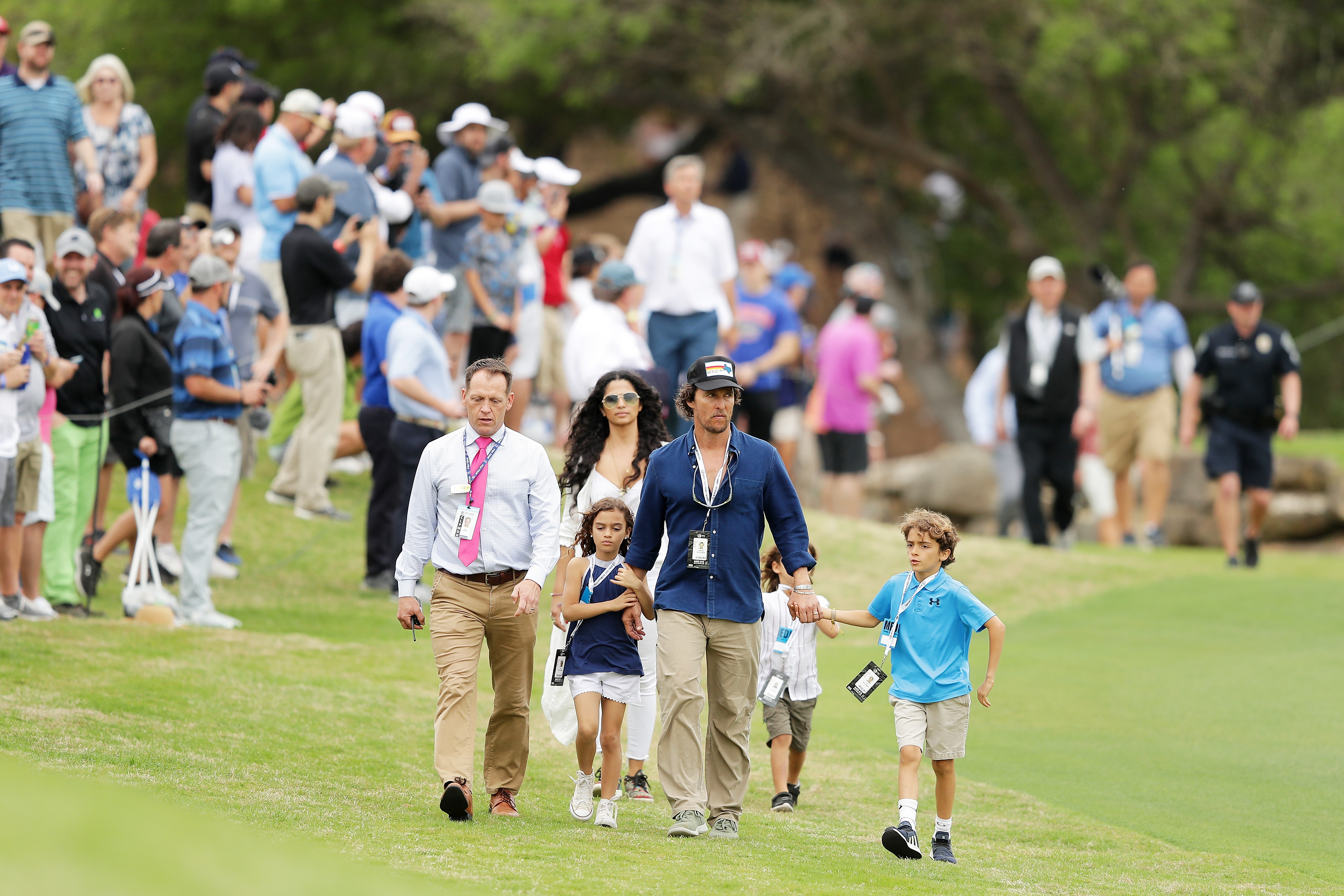 Levi McConaughey and Vida McConaughey walk beside Matthew McConaughey across the golf course grass. Levi wears a blue top and shorts, while Vida walks close in a light outfit with sneakers. Livingston McConaughey trails slightly behind as Camila Alves walks alongside the group.