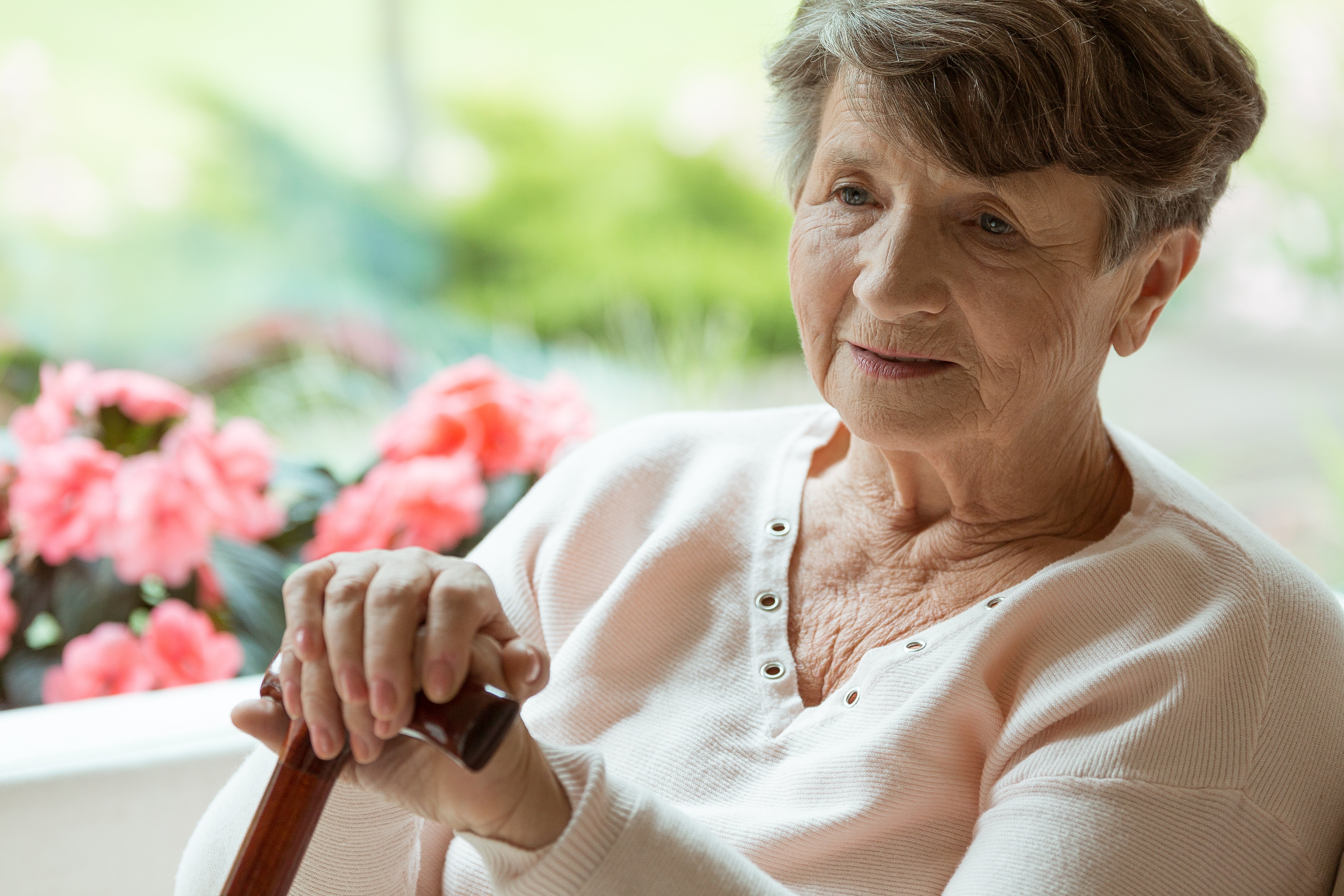 Elder woman sitting on white sofa with walking cane in room with pink flowers | Photo: Shutterstock.com