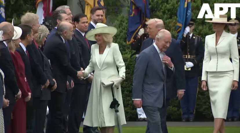 As Queen Camilla continues down the line, shaking hands with officials, King Charles III moves ahead at a measured pace, while U.S. President Donald Trump appears just behind them, closing the gap after stepping into the sequence. | Source: YouTube/APT