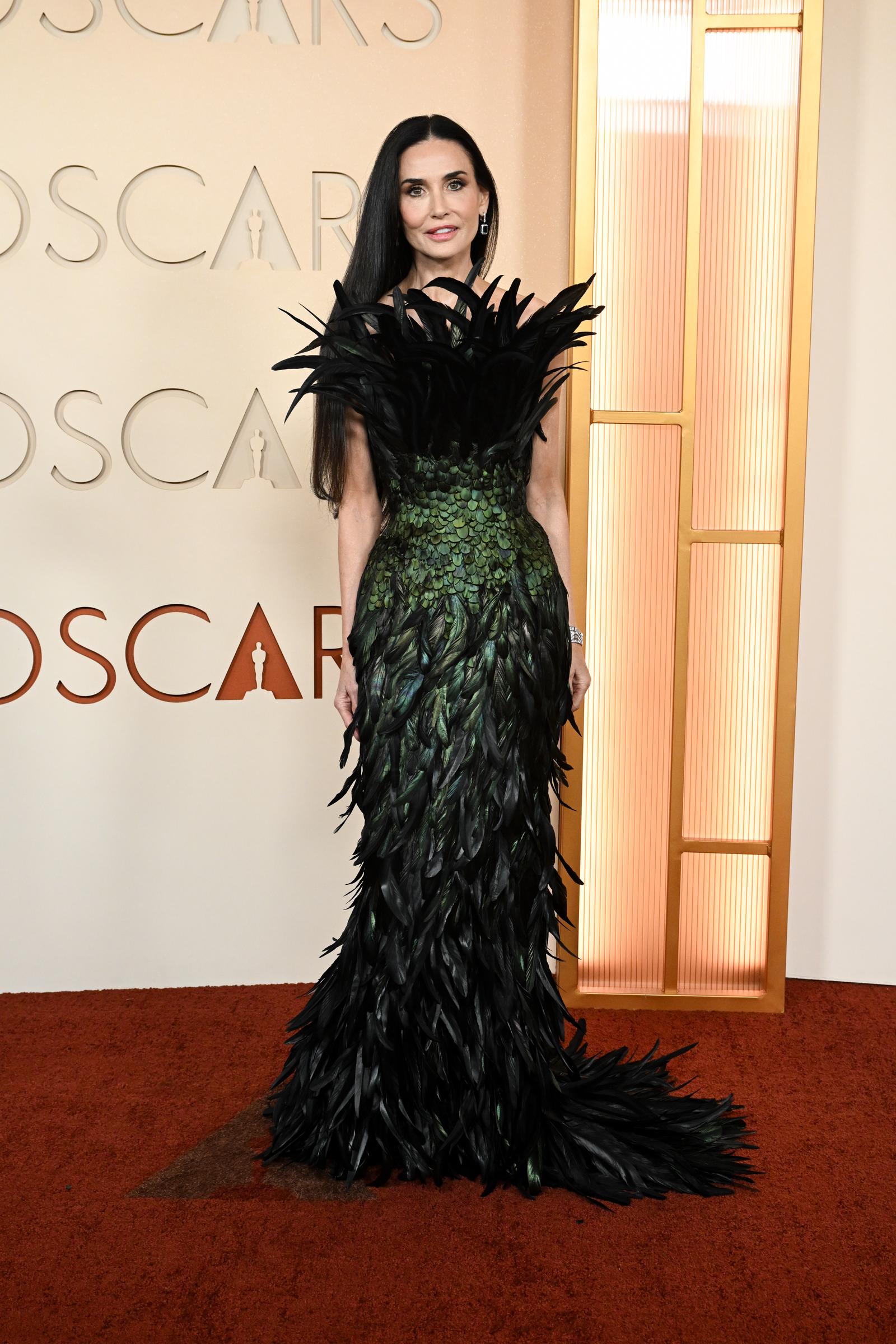 Demi Moore arrives at the 98th Academy Awards in a floor-length dark green gown constructed entirely of iridescent feathers, with a dramatic oversized feathered collar fanning out at the shoulders and a feathered train extending behind her.