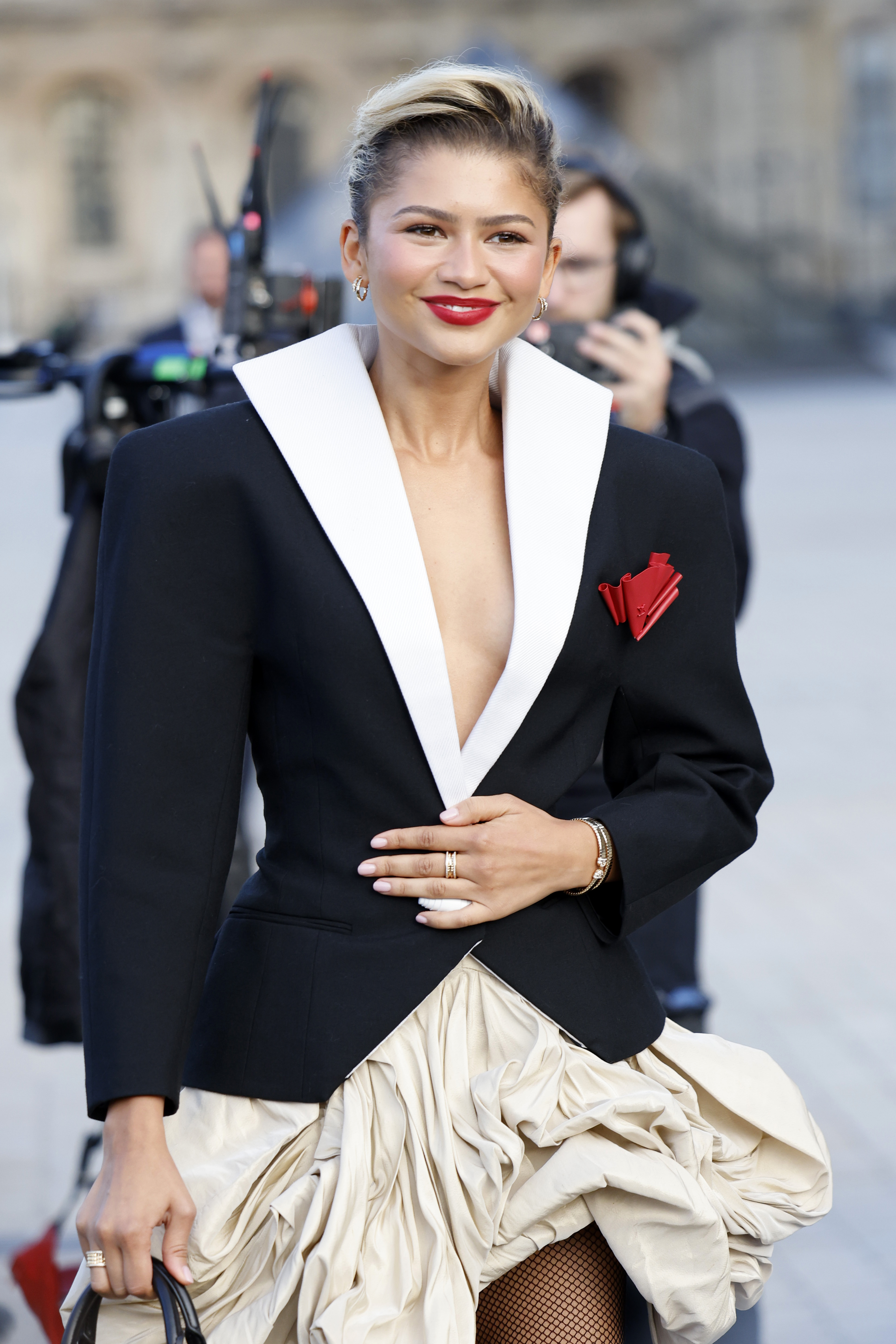 Zendaya attended the Women’s Spring/Summer 2025 show at Paris Fashion Week in France, wearing a sculpted black plunging blazer with exaggerated shoulders and a high white collar. The blazer, fastened with a single white cylindrical button, featured a red rose-like appliqué. She layered it over a beige bubble skirt, completing a bold, avant-garde look.