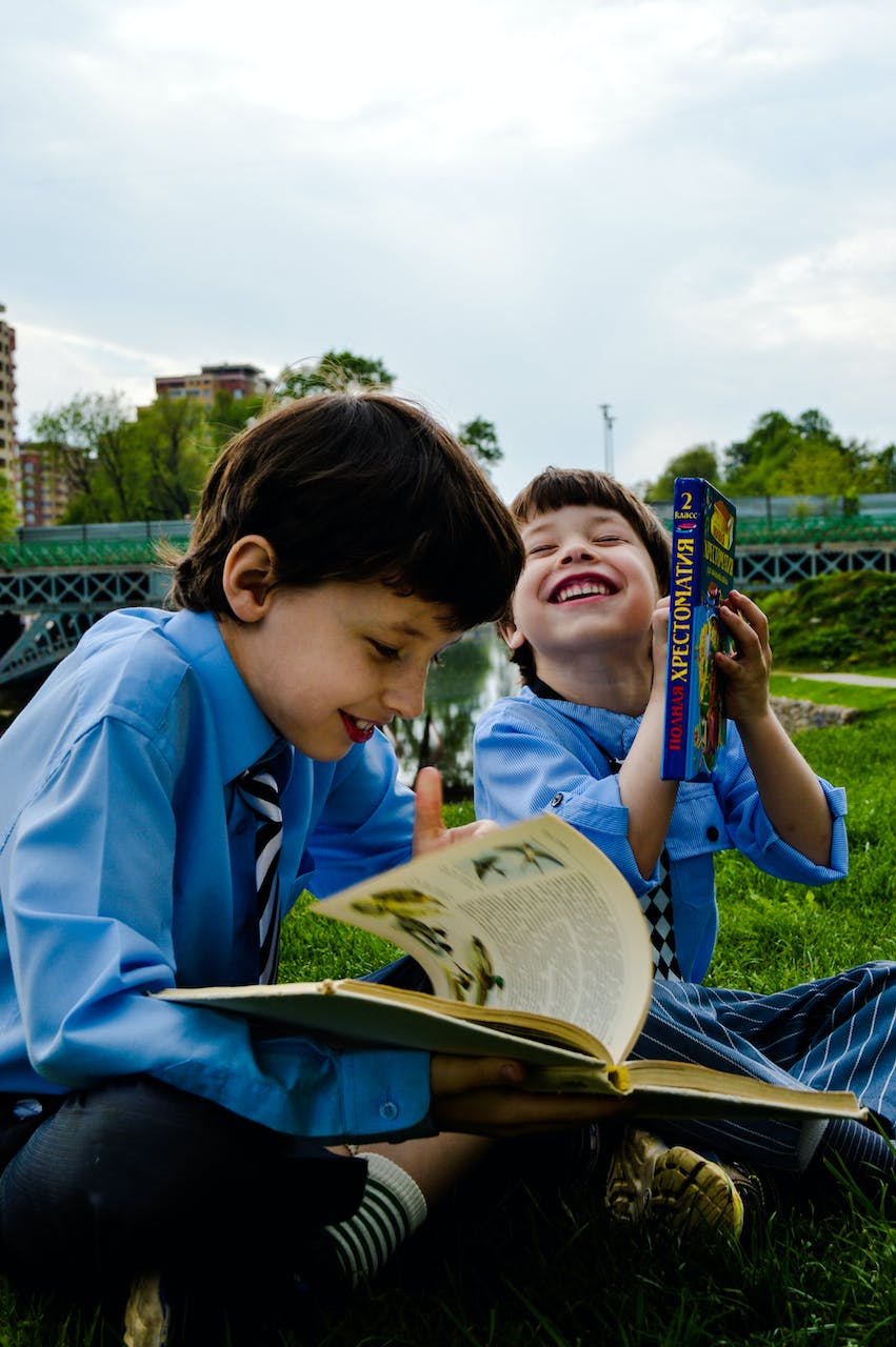 Two boys seated on the grass while laughing and reading books | Source: Pexels