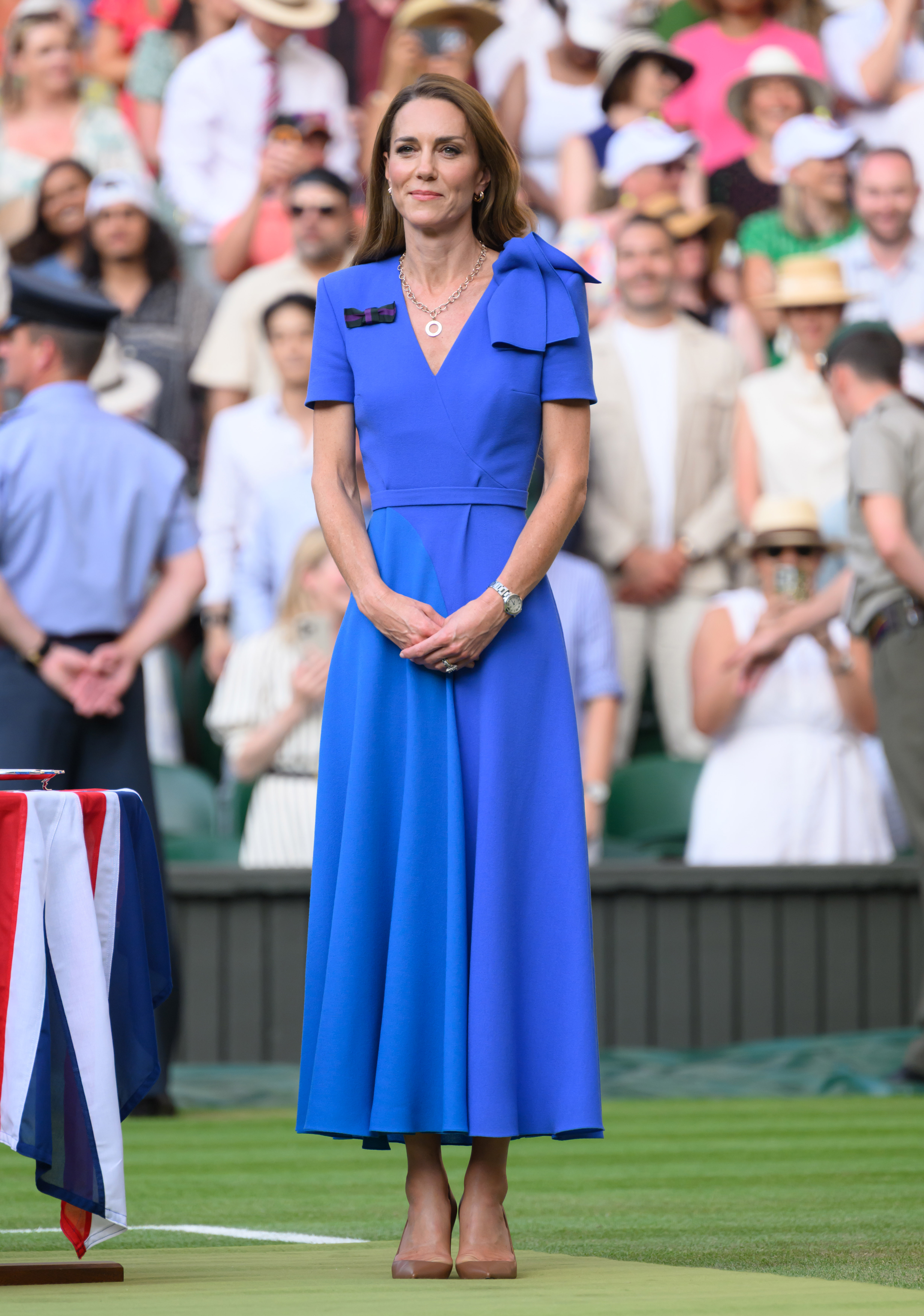 Princess Catherine stands on Centre Court in her vibrant blue dress during the Wimbledon event. Her hands are gently clasped as she faces the crowd.