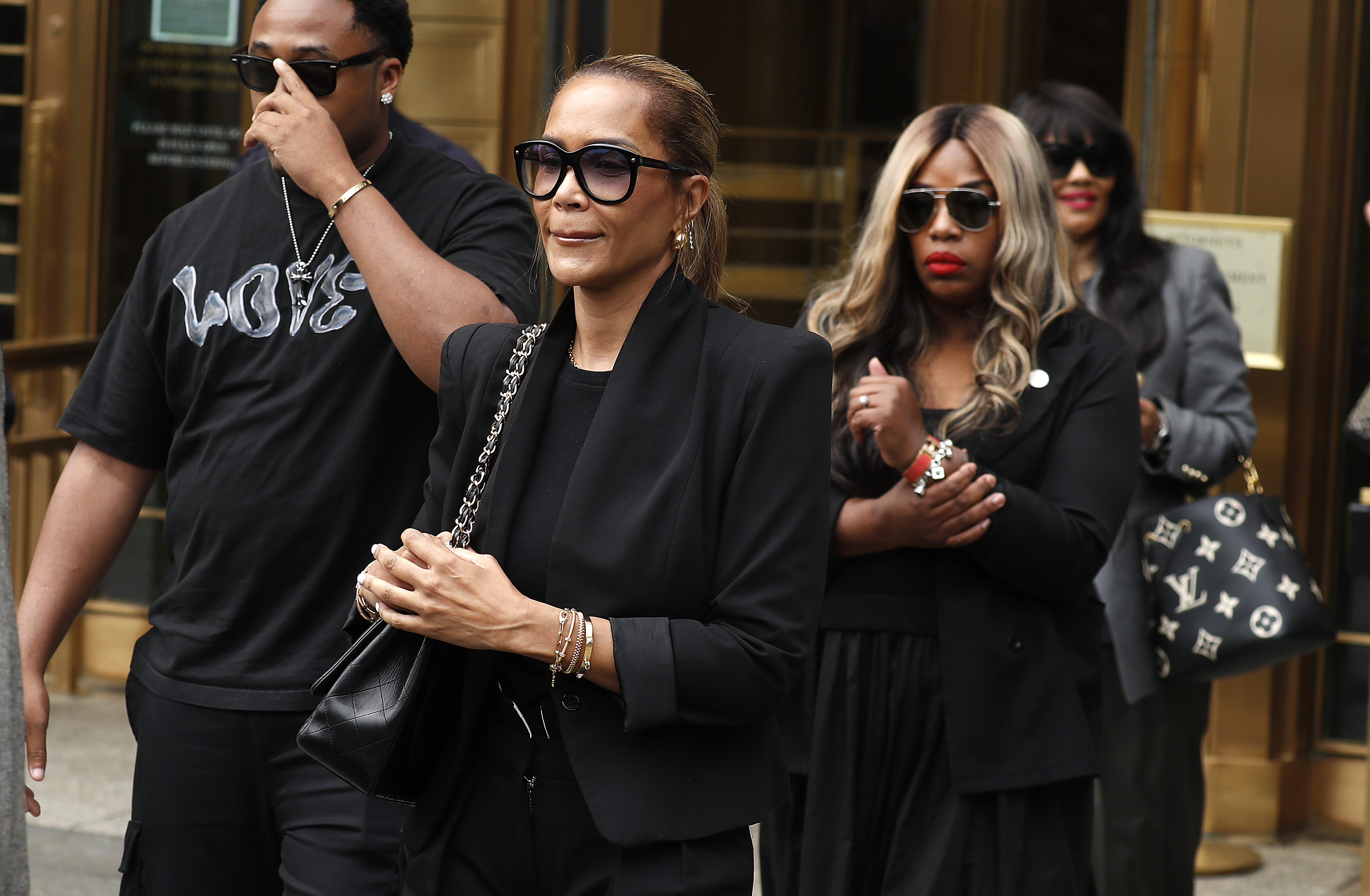 Sarah Chapman exits the Manhattan court with a composed expression, wearing black sunglasses. The mood around her is tense, as others follow closely without speaking.