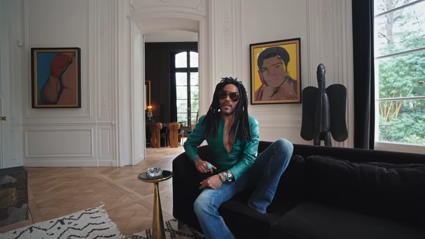 Lenny lounges on a plush black sofa in front of an iconic Andy Warhol portrait of Muhammad Ali and a dark, winged African sculpture. | Source: YouTube/@Archdigest