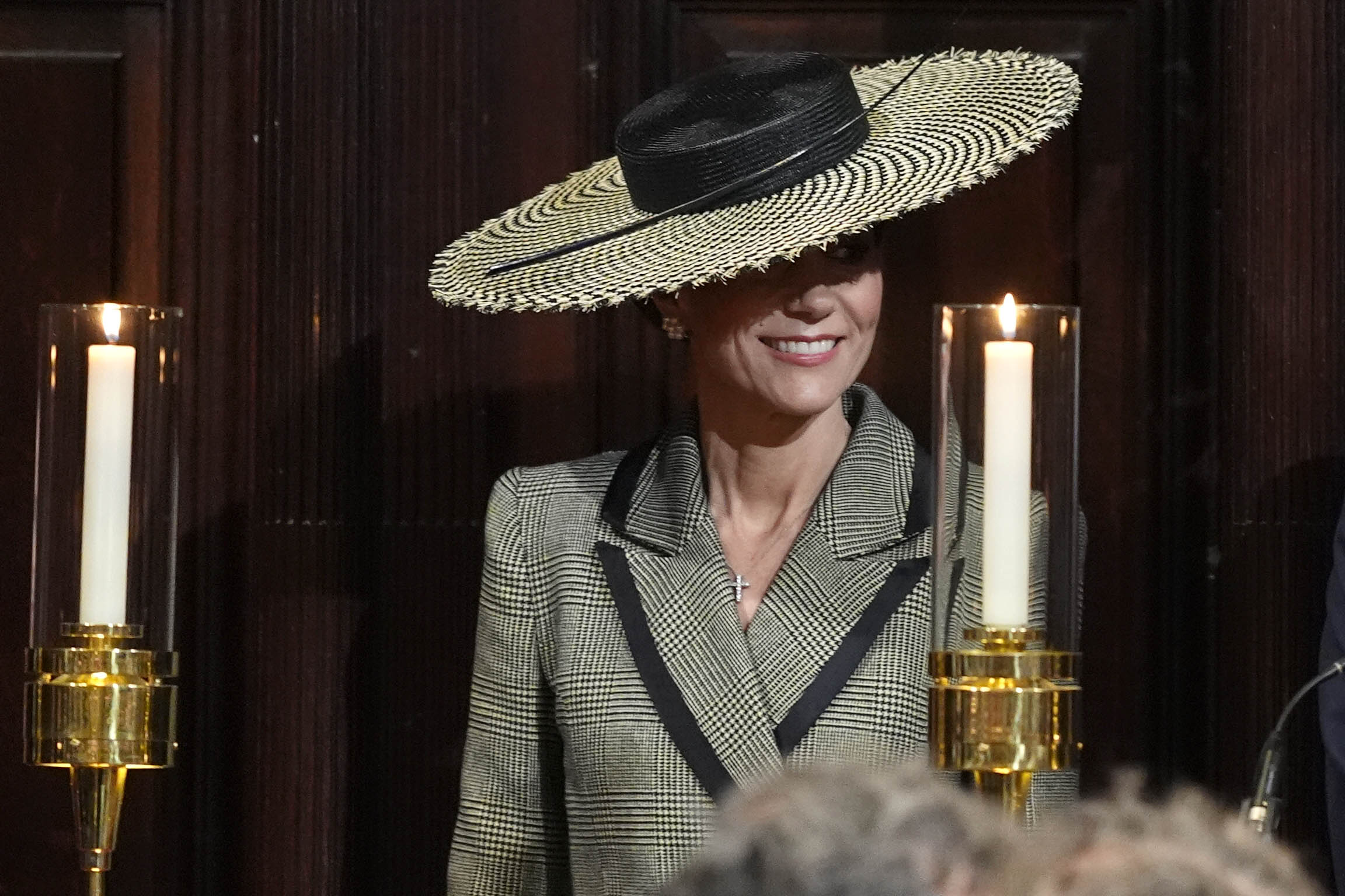 Inside Canterbury Cathedral, the Princess of Wales is flanked by two tall candlesticks, their flames casting a warm glow against the dark wood panelling behind her. The wide houndstooth brim of the Juliette Botterill Millinery boater frames her face almost theatrically in this setting, and the diamond cross necklace is clearly visible at her neckline — a considered choice for a ceremony marking a historic moment in the Church of England's history.