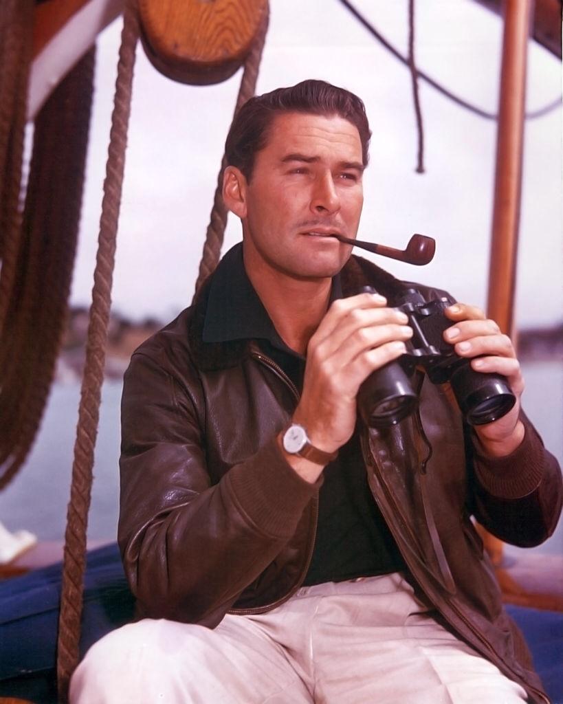 Australian actor, wearing a brown leather jacket and smoking a pipe while holding a pair of binoculars, 1950 | Photo: Getty Images