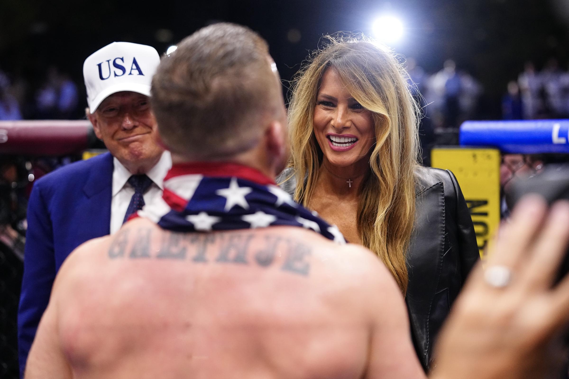 Melania Trump smiles broadly while greeting attendees, revealing a more animated expression. Bright arena lighting highlights her contoured features and polished appearance.