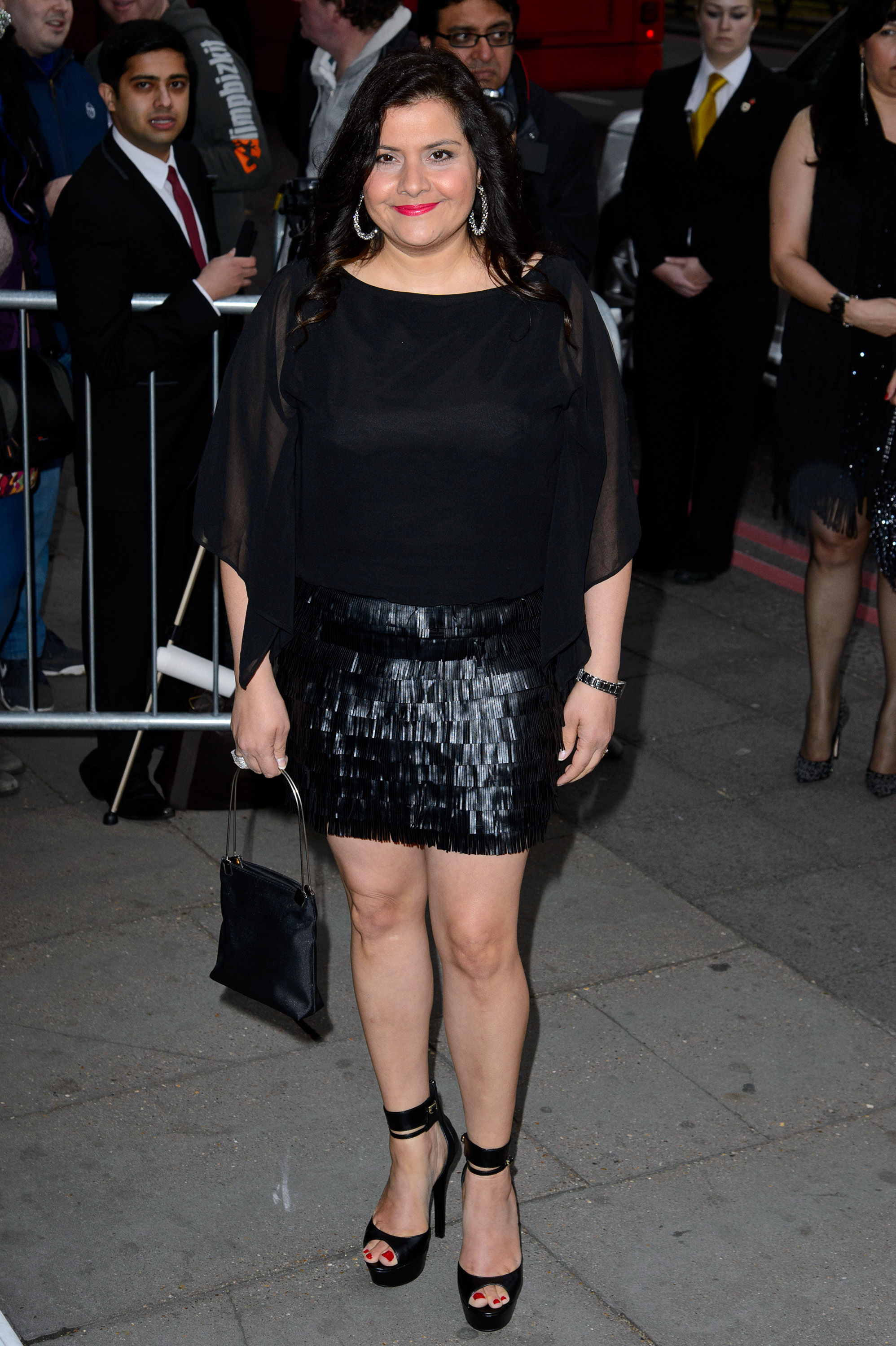 Wadia arrives at The Asian Awards at the Grosvenor House Hotel in April 2014 looking noticeably more confident in a bold all-black ensemble — a sheer floaty blouse with split sleeves over a tiered black leather-look fringe mini skirt. She pairs the statement look with strappy black platform heels, a small structured black handbag, and a bright slash of red lipstick.