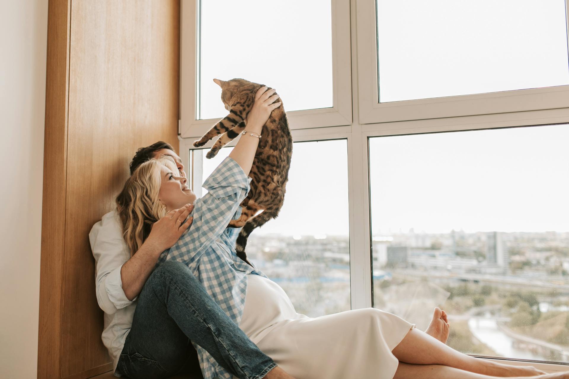 A pregnant couple sitting by the window and playing with their cat | Source: Pexels