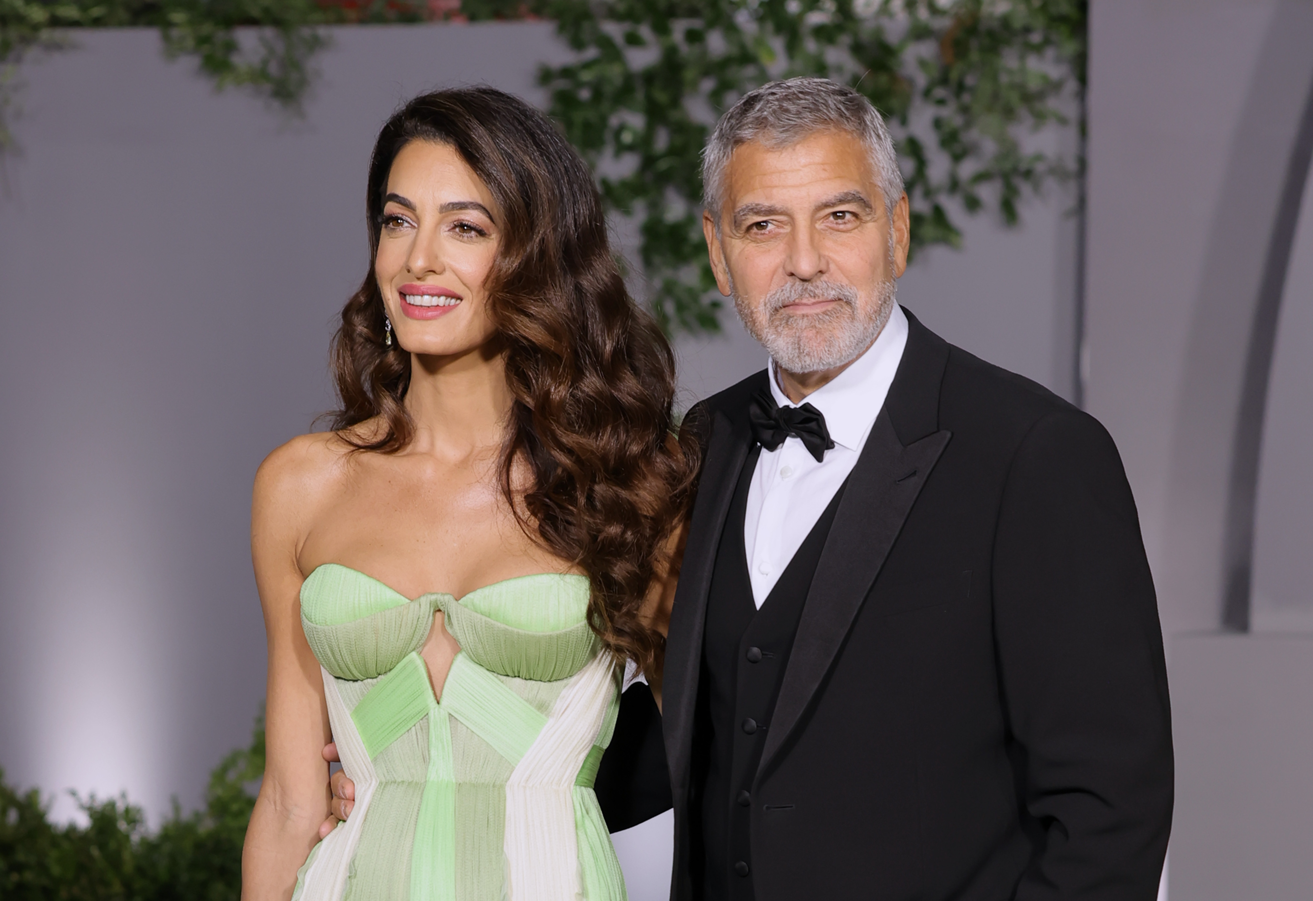 At the Academy Museum Gala, Amal wears a strapless pastel green and white gown featuring a structured bodice and keyhole detail, while George Clooney stands beside her in a classic black tuxedo with a bow tie and vest.