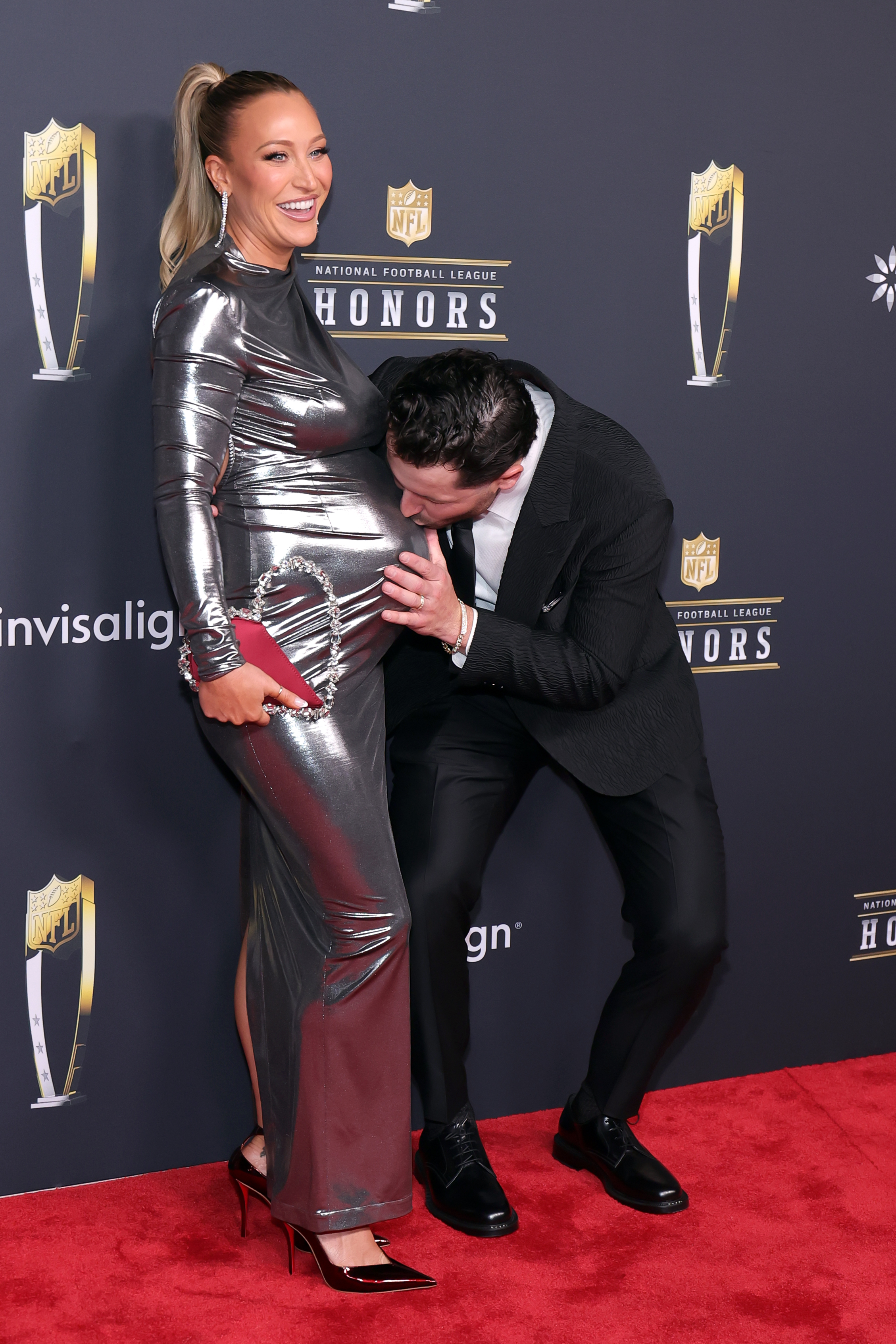 Emily Wilkinson laughs as Baker Mayfield affectionately kisses her baby bump, the couple sharing a playful, intimate moment that contrasts with the evening's formal tone.