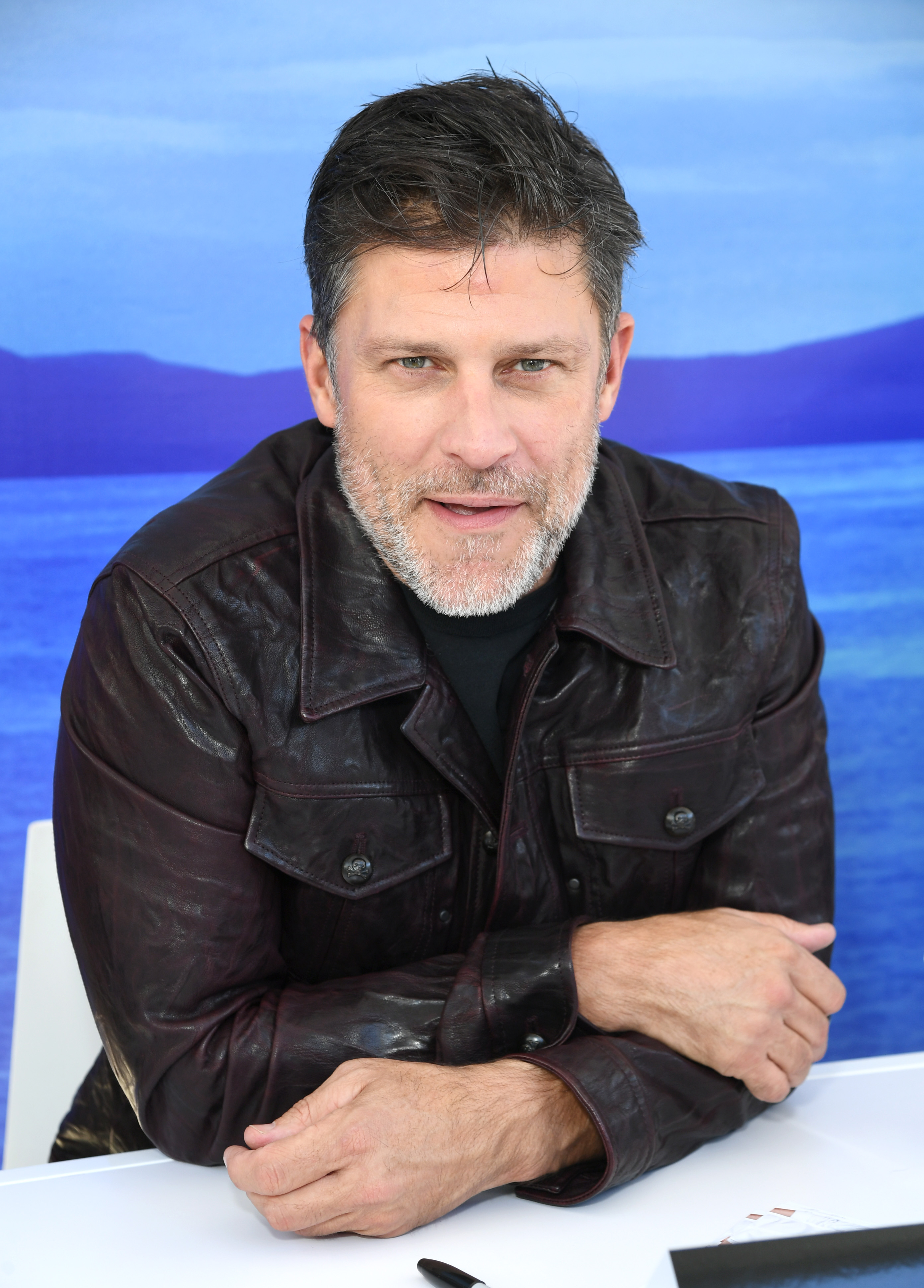 Greg Vaughan is seen at a fan event in 2022, seated at a signing table in front of a scenic blue backdrop. Wearing a deep burgundy leather jacket over a black shirt, he looks directly at the camera with a friendly, relaxed expression. His salt-and-pepper beard adds a distinguished touch to the candid portrait.