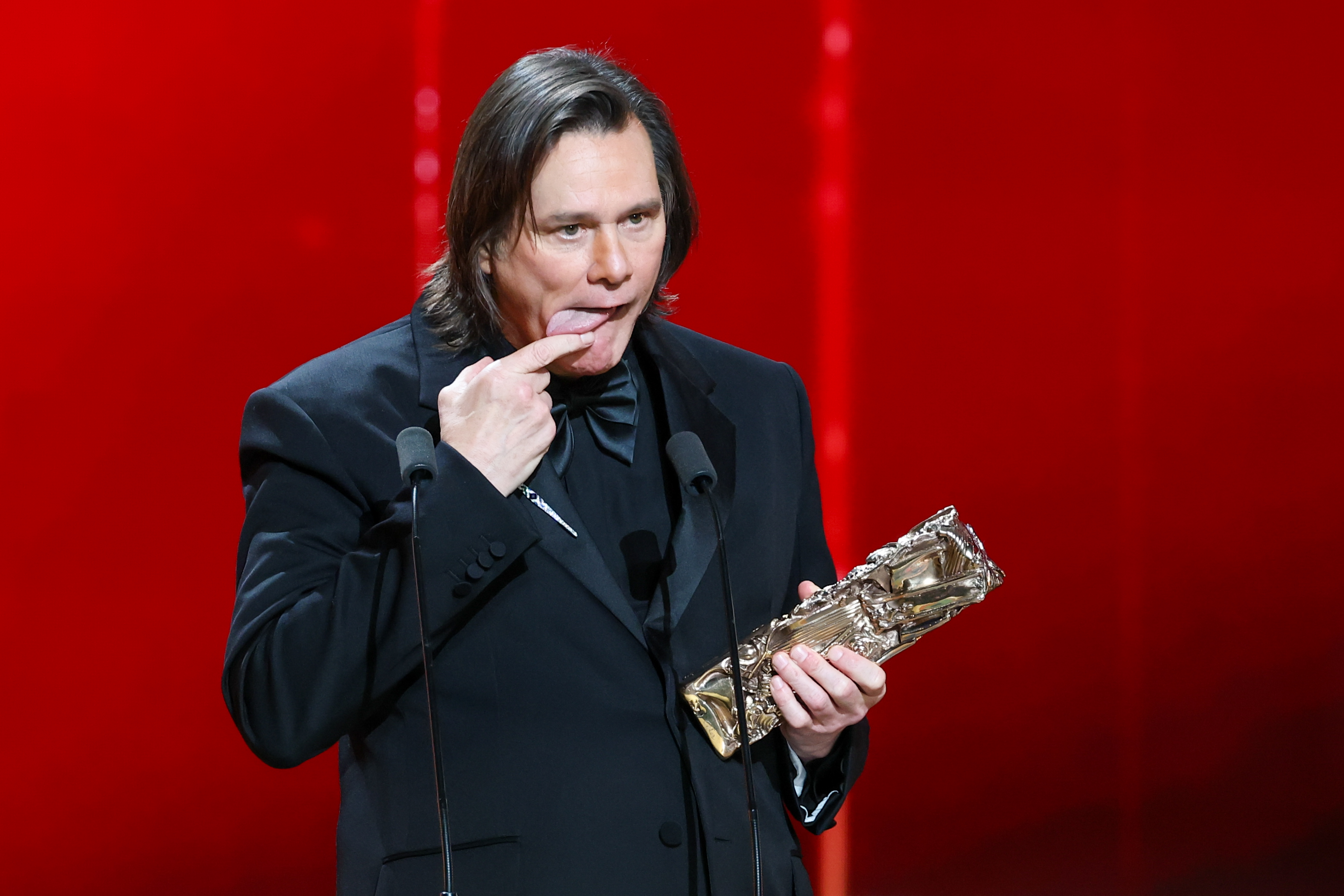 Holding his Honorary César Award, Jim Carrey playfully points to his tongue mid-speech. Dressed in a classic black tuxedo, he stands between two microphones against a striking red backdrop. The light hearted moment unfolds onstage at the 51st César Film Awards at L'Olympia in Paris, France.
