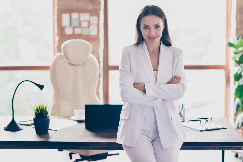 Mujer recostada de un escritorio luciendo un blazer blanco con pantalones blancos de vestir. | Foto: Shutterstock Mujer recostada de un escritorio luciendo un blazer blanco con pantalones blancos de vestir. | Foto: Shutterstock
