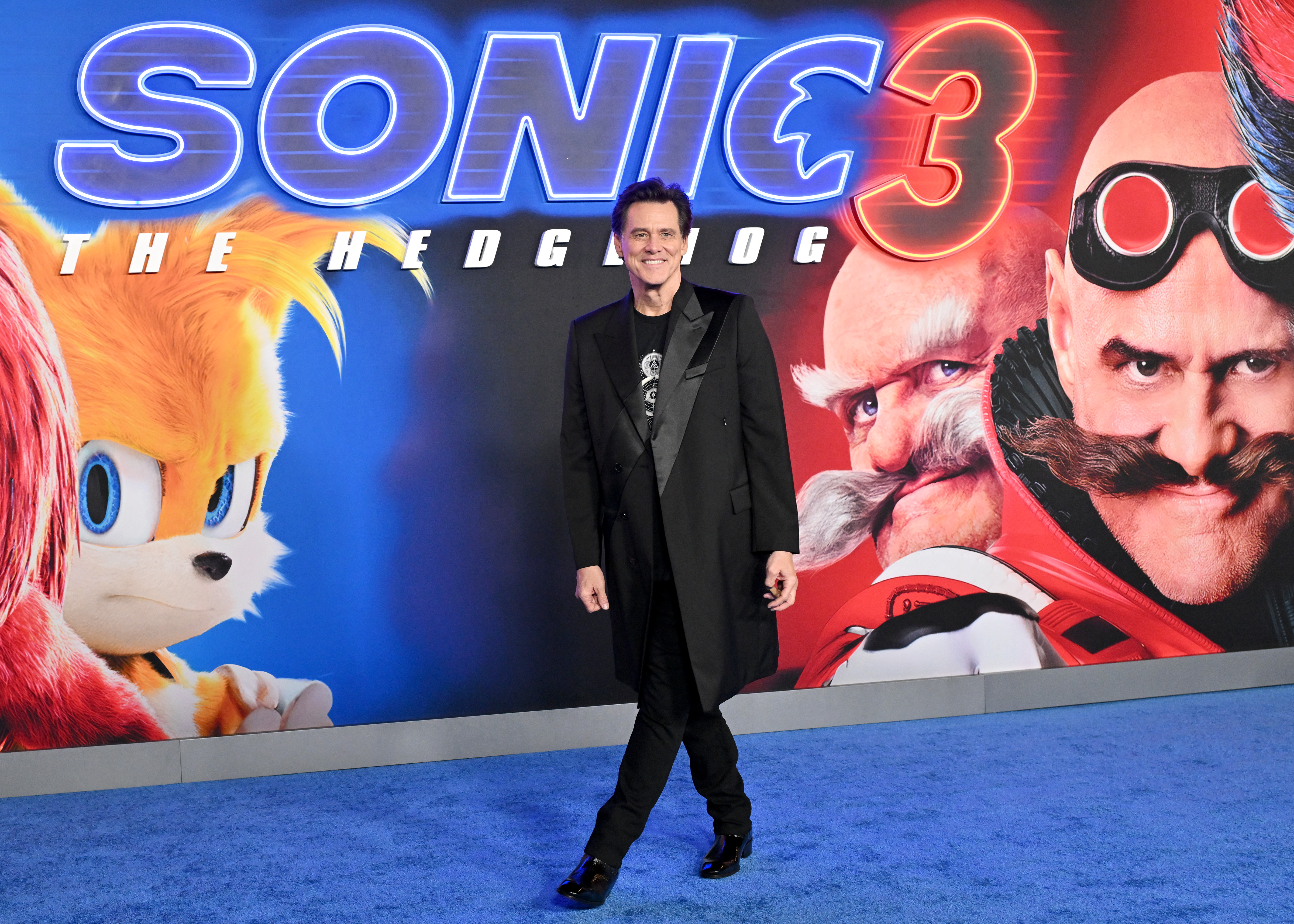 Jim Carrey attends the Los Angeles premiere of Paramount's "Sonic the Hedgehog 3" at the TCL Chinese Theater. Dressed in an elegant black coat and matching pants, he poses confidently on the blue carpet. The vibrant backdrop featuring Sonic characters highlights his iconic role in the beloved film franchise.