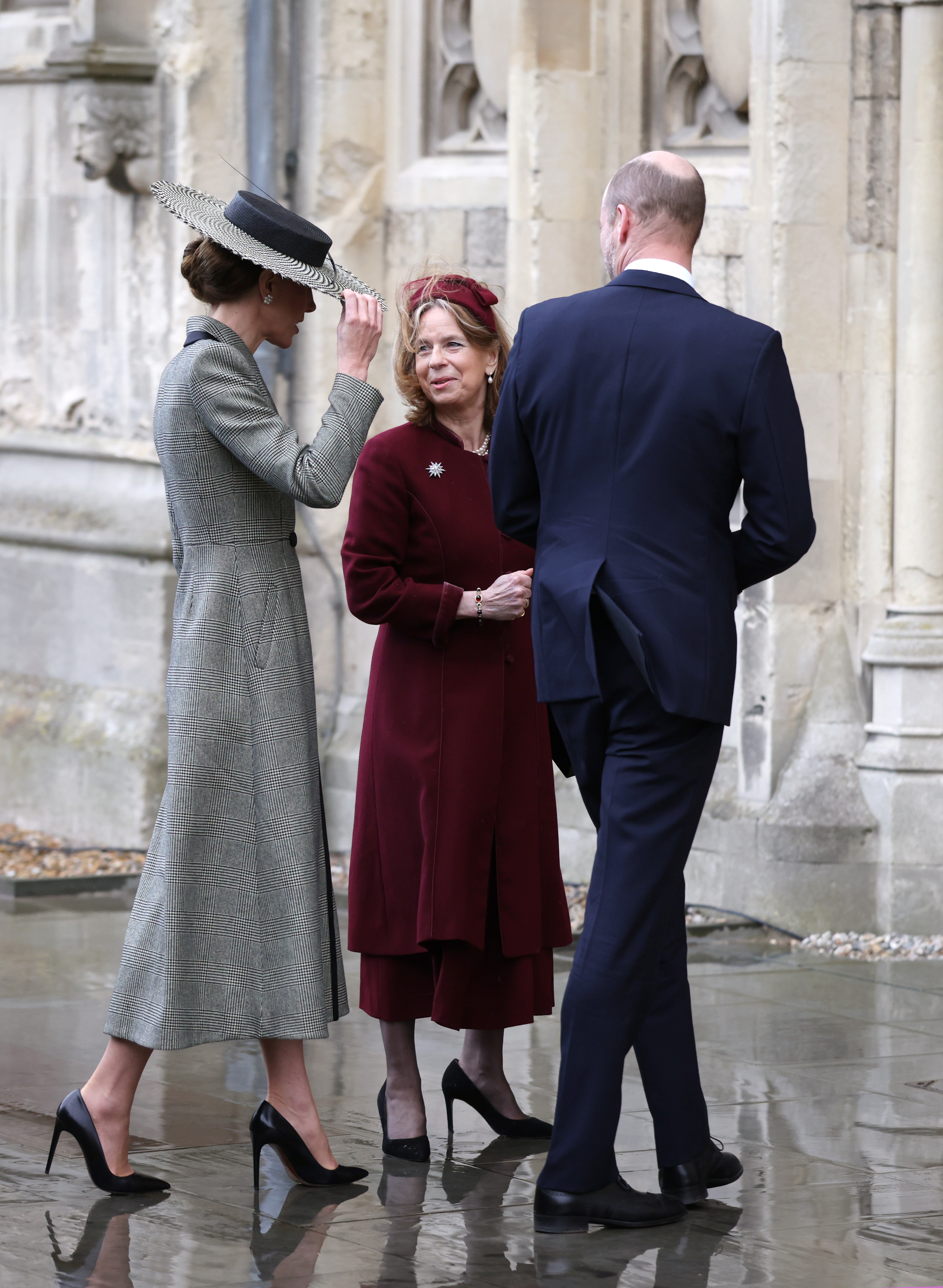 One hand up, hat firmly gripped — the Princess of Wales gets her first real taste of the Canterbury wind as she pauses to chat outside the cathedral's ancient stone walls. Prince William, in a navy suit, stands beside her with his back to the camera, seemingly unaffected, while the wide houndstooth brim tilts at a precarious angle that told the whole story before the internet even got to it.