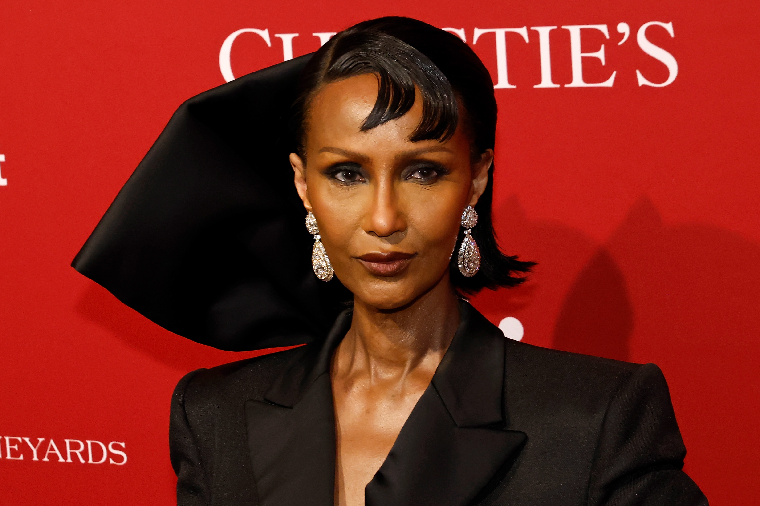 A close-up reveals the full impact of Iman's beauty look: smoky, heavily lined eyes, a deep brown lip, and long crystal drop earrings catching the light beneath the sculptural black satin bow rising behind her head.