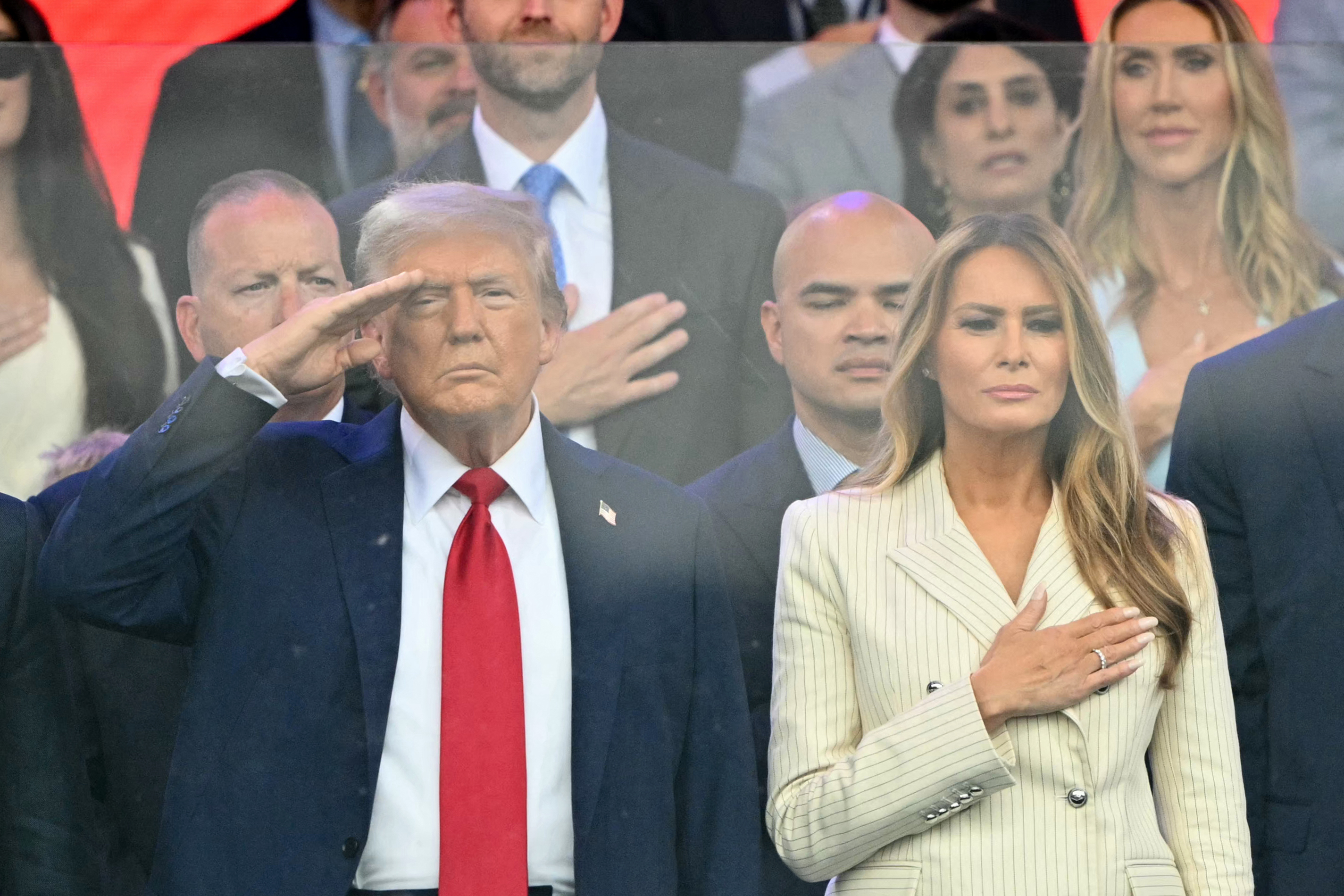Donald Trump salutes while Melania Trump places her hand on her chest during a ceremonial segment of the parade. Surrounded by officials and attendees, their synchronized gesture highlights their role as central figures in the patriotic observance.