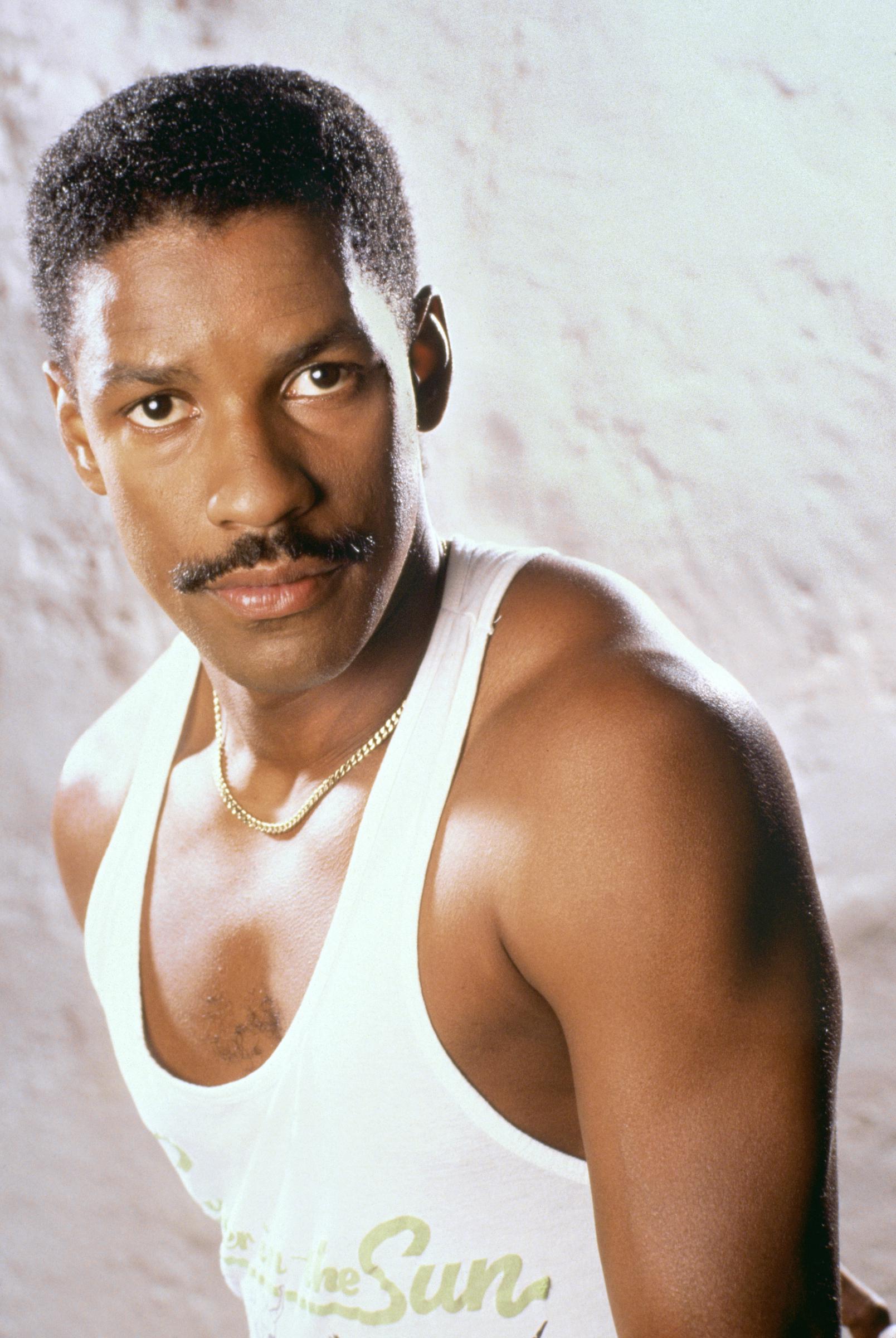 Denzel Washington exudes confidence in a white tank and gold chain in this portrait. With shortly styled hair and a mustache, his hair is sharp and polished — a style that became one of his most recognizable early looks.