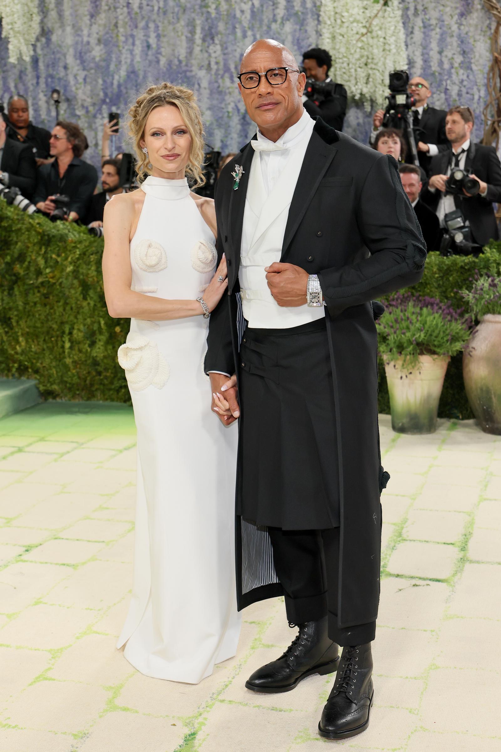 Lauren Hashian wears a sleek white gown with sculpted detailing and soft curls. The look is clean, minimal, and refined. Dwayne Johnson complements her in a structured black coat, white waistcoat, and polished boots.