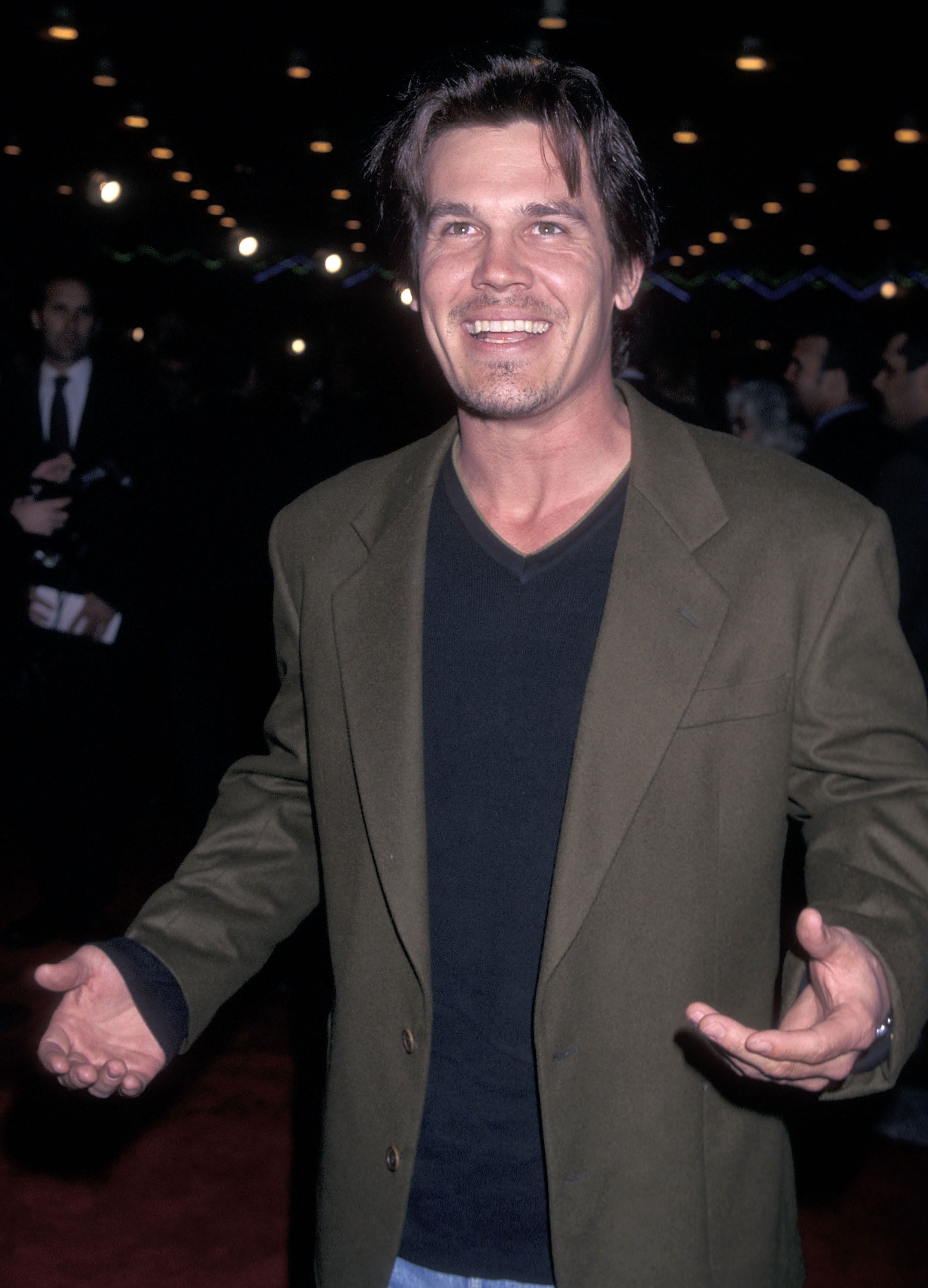 Brolin at the "Primary Colors" premiere in Universal City. He wears a deep V-neck black shirt layered under an olive green blazer, smiling with both arms raised toward the camera.