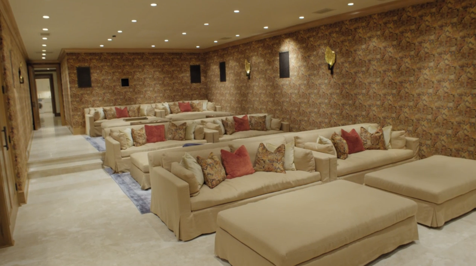 Tiered rows of plush neutral-toned sofas and oversized ottomans provide comfortable seating throughout this custom home theater. Patterned wallpaper, warm sconce lighting, and built-in wooden cabinetry featuring a wall-mounted flat-screen TV give the media room a cozy, inviting atmosphere. | Source: YouTube/Architectural Digest