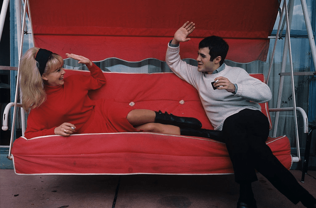 Sophie DAUMIER, vêtue d'un pull à col roulé rouge, un bandeau noir dans ses cheveux blonds et Guy BEDOS, pull blanc sur pantalon noir s'amusent, cigarettes en main et bras levés, l'une allongée, l'autre assis, sur une balancelle de tissu rouge. | Photo : Getty Images Sophie DAUMIER, vêtue d'un pull à col roulé rouge, un bandeau noir dans ses cheveux blonds et Guy BEDOS, pull blanc sur pantalon noir s'amusent, cigarettes en main et bras levés, l'une allongée, l'autre assis, sur une balancelle de tissu rouge. | Photo : Getty Images