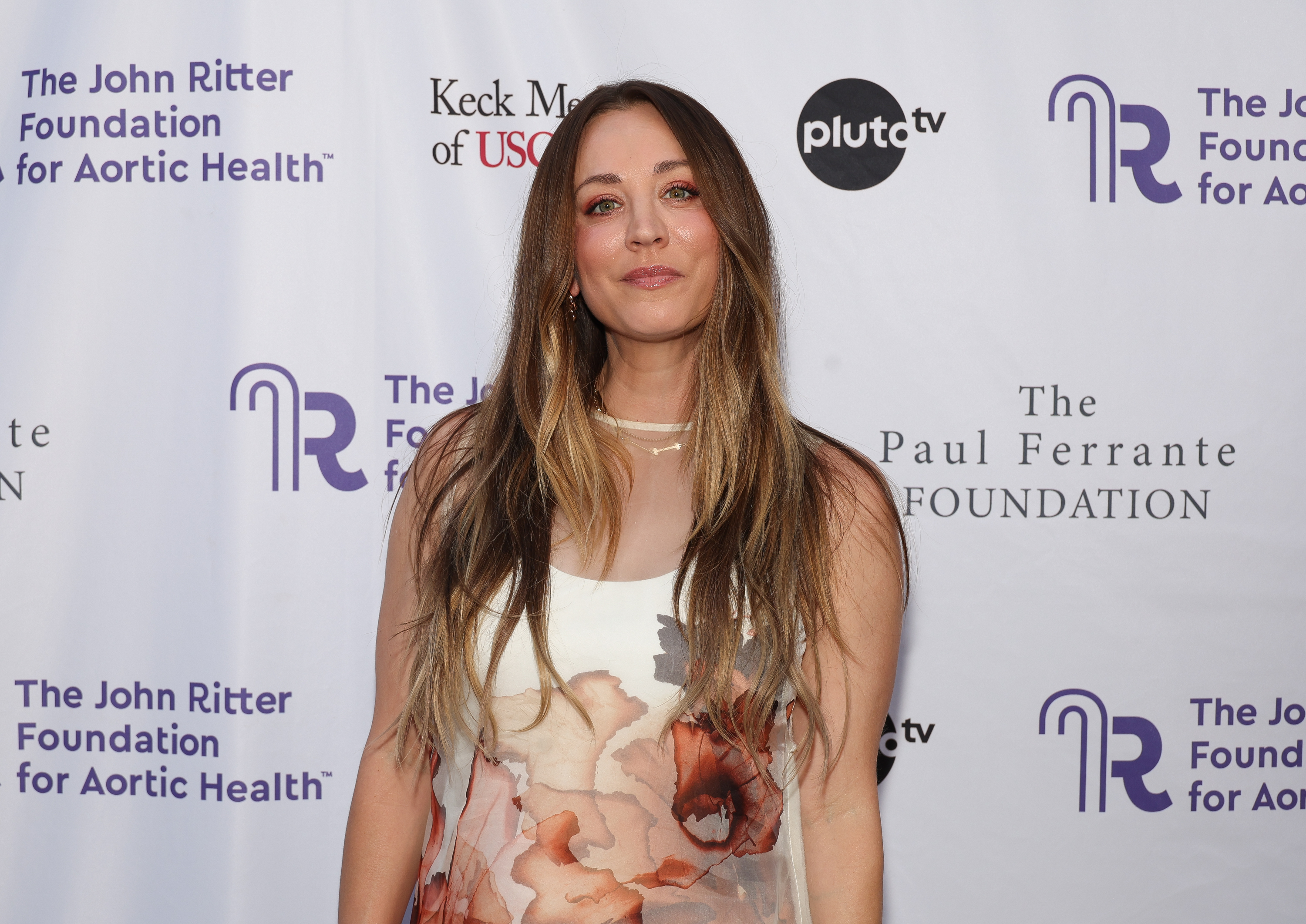 Kaley Cuoco attends the John Ritter Foundation event in a sheer floral-print dress with soft pink and red tones. Her long, wavy hair and minimal jewelry give the look a relaxed, elegant vibe.