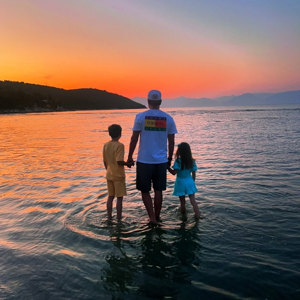 Posted on 11 August 2024, Adam Thomas is seen standing ankle-deep in calm waters at sunset, holding hands with his two children as they face the horizon together — a peaceful and reflective family moment that highlights his role as a devoted father beyond the spotlight. | Source: Instagram/carolinethomas23