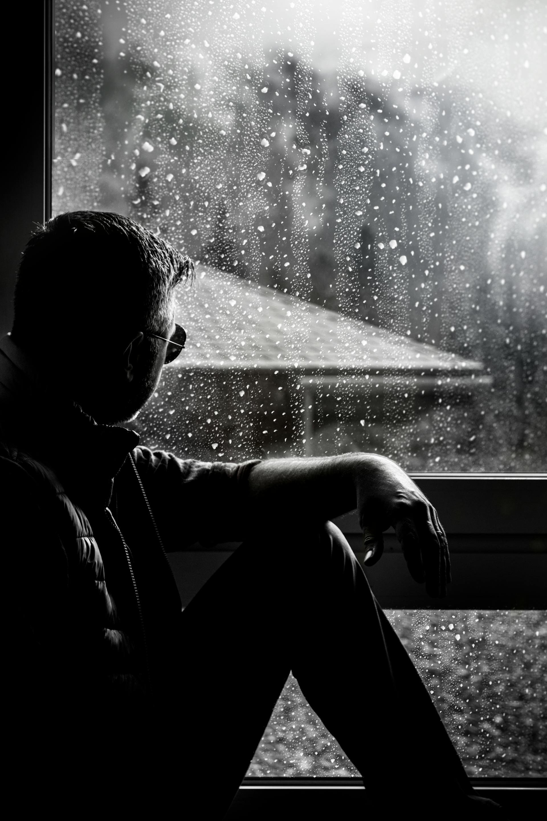A grayscale photo of a man sitting beside a window covered with raindrops | Source: Pexels
