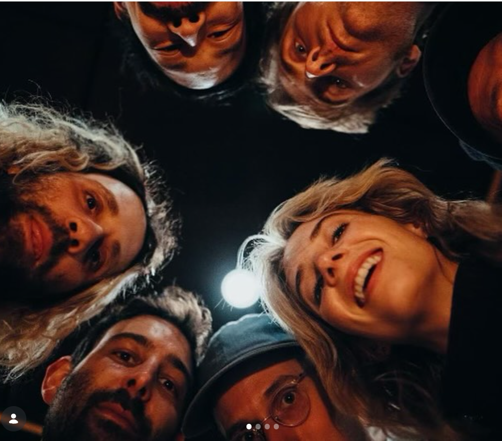A group of friends huddle in a circle, looking down at the camera as a bright light glows between them, with Maya Hawke smiling warmly among the group. | Source: Instagram/maya_hawke