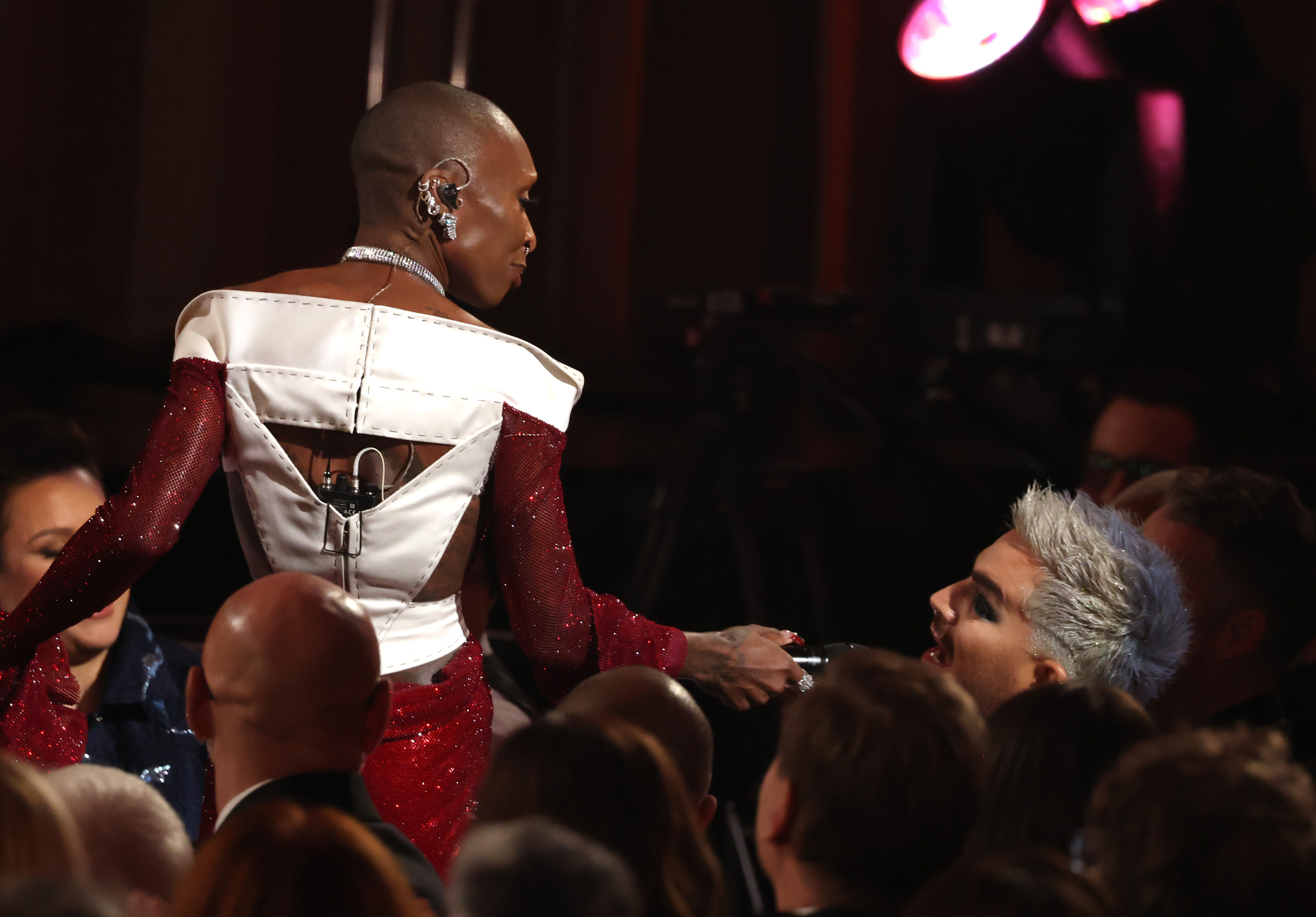 Cynthia Erivo hands the microphone to Adam Lambert, who appears to launch into a spontaneous vocal moment. Her structured red-and-white gown gleams under the lights, while Lambert’s signature silver hair rises above the crowd in a striking contrast.