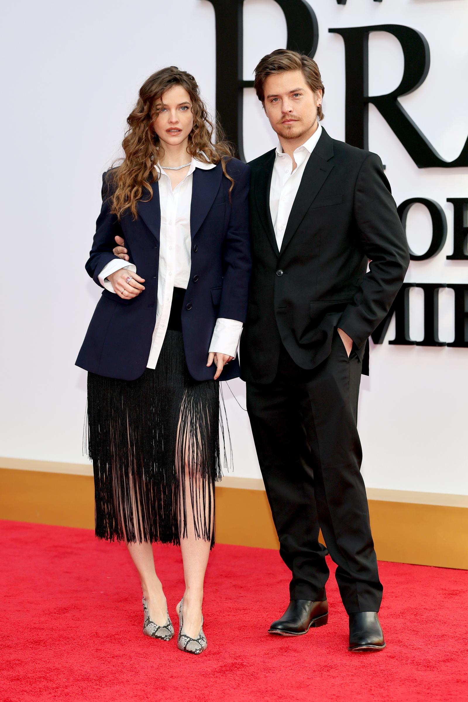 Barbara Sprouse wears a navy oversized blazer over a white collared shirt and a black fringe midi skirt, finished with snakeskin-print kitten heels and a delicate diamond necklace, while her husband Dylan opts for a classic black suit with a white open-collar shirt and black Chelsea boots.