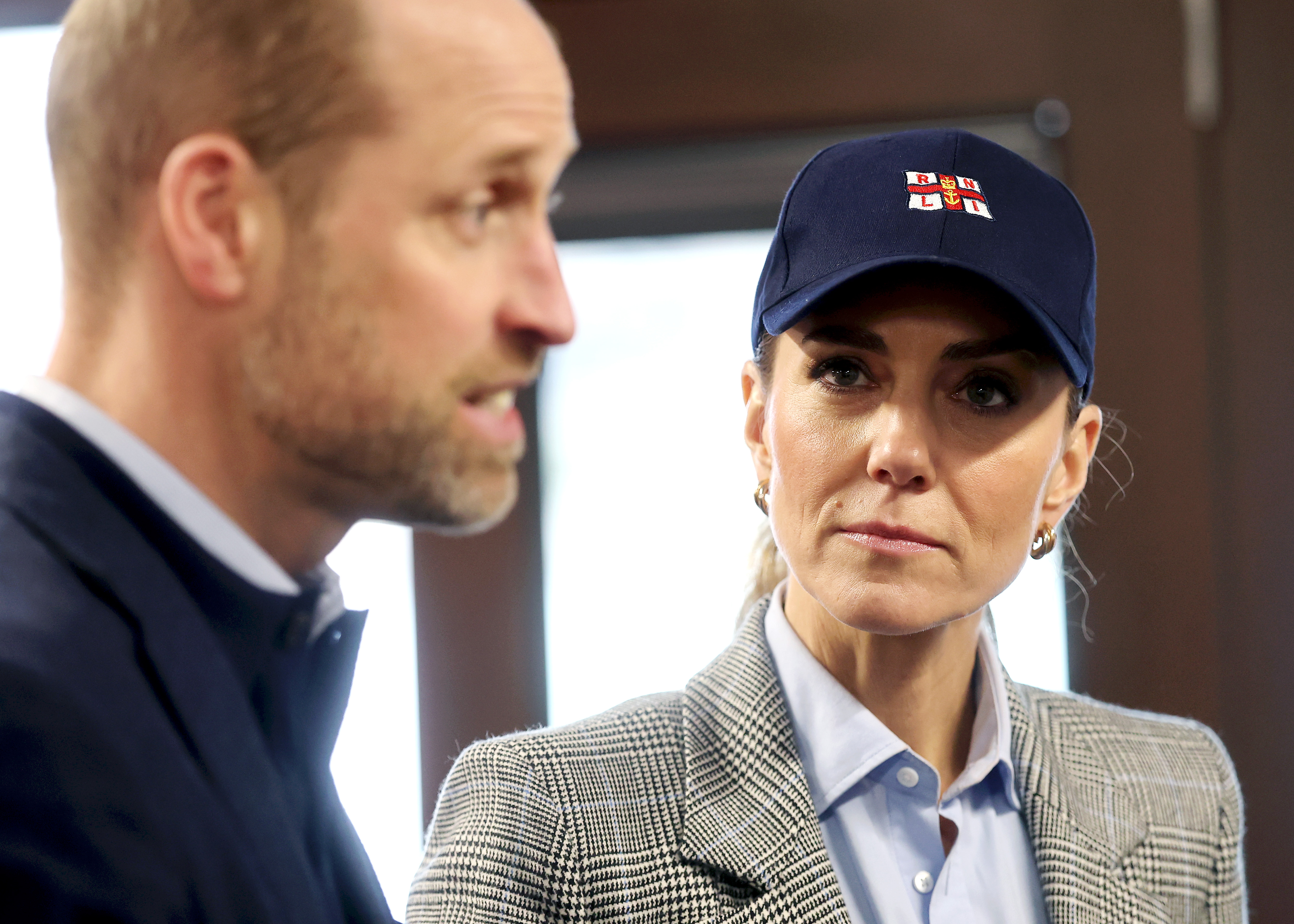 The Princess of Wales listens intently, her gaze fixed on the Prince of Wales mid-conversation, during their visit to the RNLI Tower Station on 12 March 2026 — a navy RNLI cap now replacing the grey blazer's more polished earlier look from the brewery. The couple were there to hear first-hand about the life-saving work carried out by staff and volunteers on the River Thames.