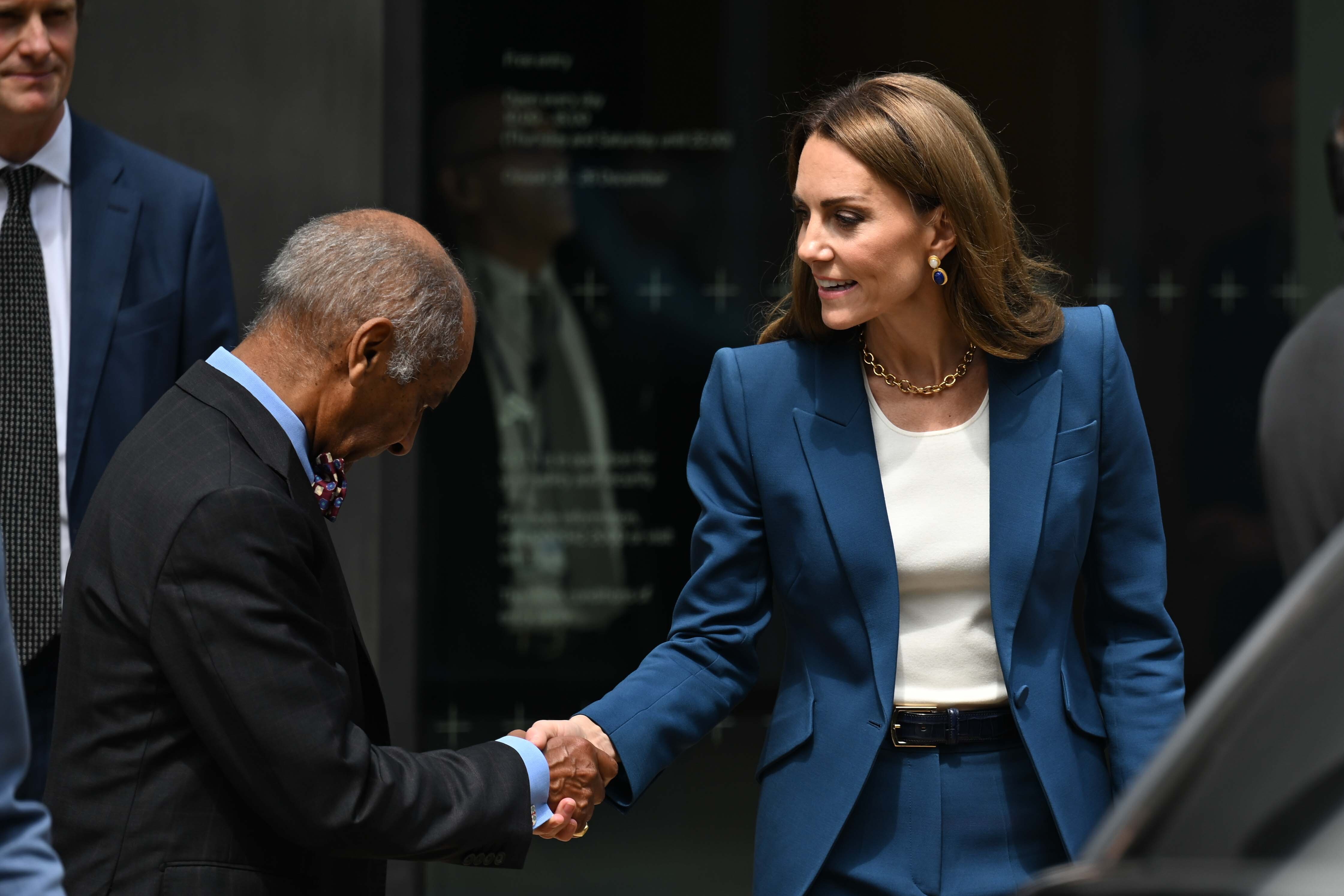 With a polite nod and steady eye contact, Princess Catherine greets an official at the V&A East Storehouse. Her posture is graceful, her smile subtle, and her sapphire earrings add a refined accent to her outfit.