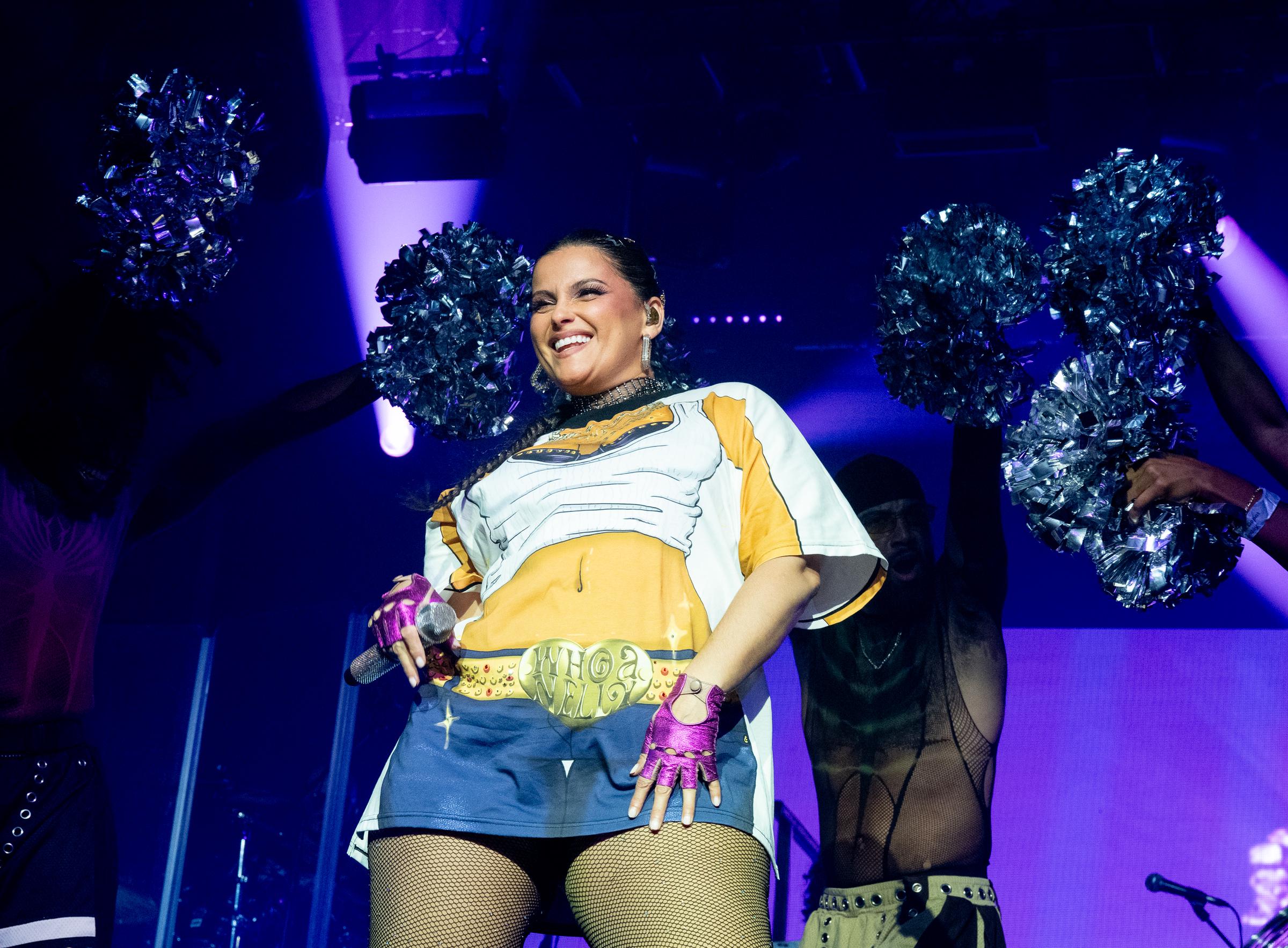 Furtado is joined by dancers holding pom-poms as she performs in a cartoon-printed oversized T-shirt and fishnet tights. She smiles while holding a microphone, wearing bright pink gloves and a bold “Whoa Nelly” belt design on her outfit.